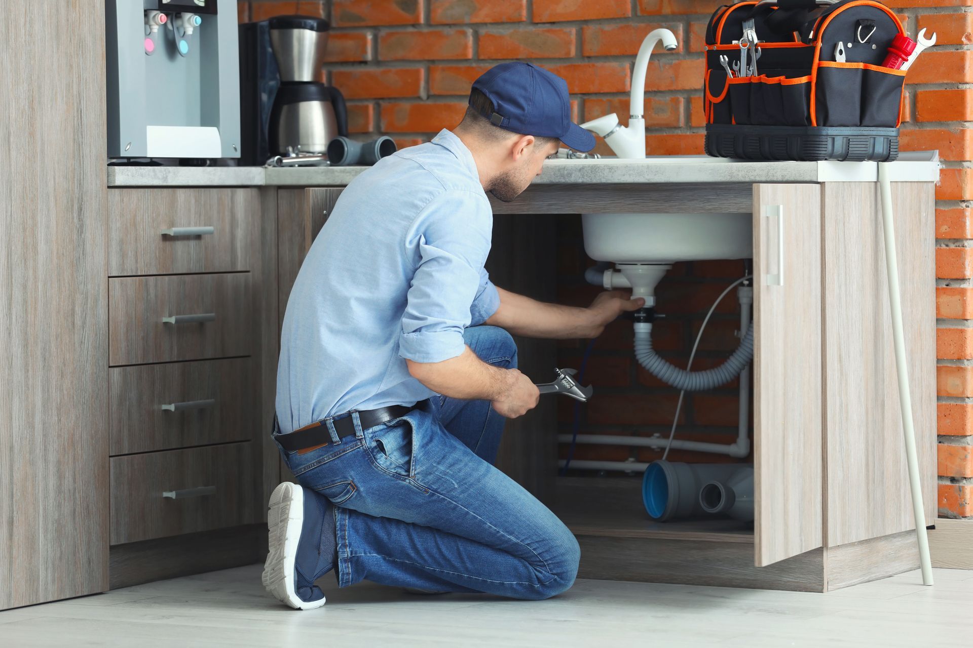 A person in a blue shirt and cap kneels in a kitchen to repair the pipes under the sink.