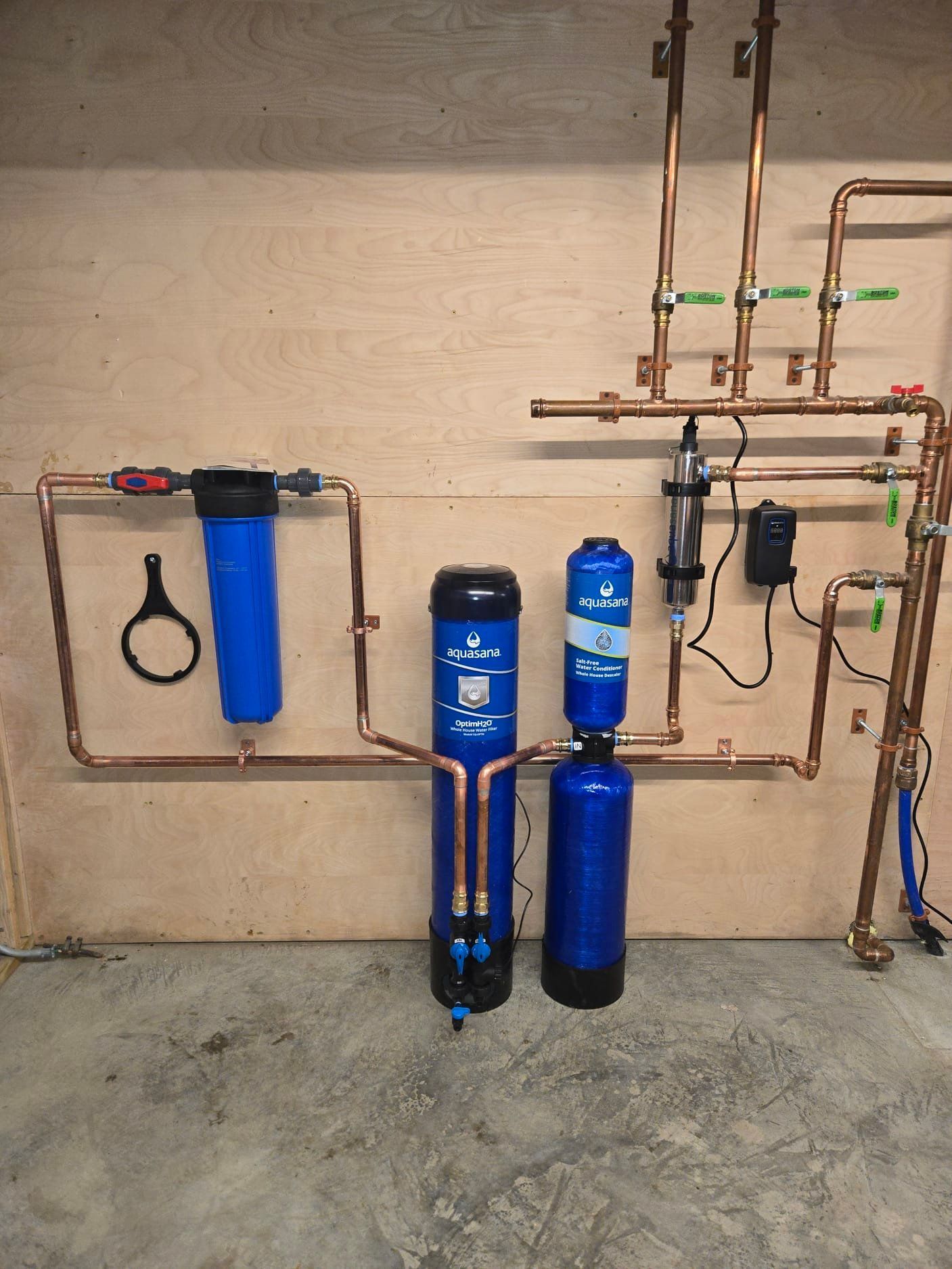 A home water filtration system with blue tanks and filters mounted on a plywood wall, connected by copper piping.