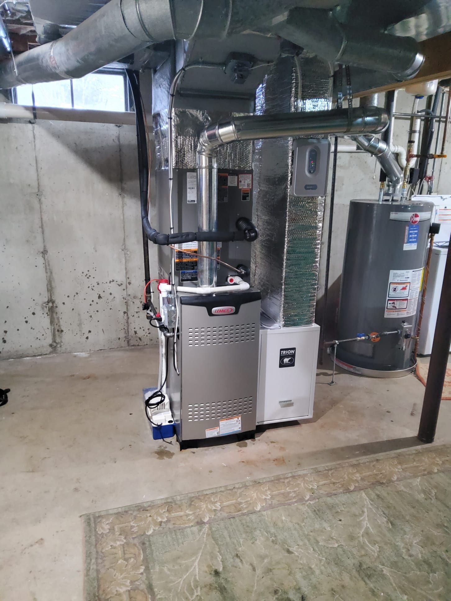 A silver furnace and white air purifier unit stand in a basement with concrete walls and a water heater nearby.