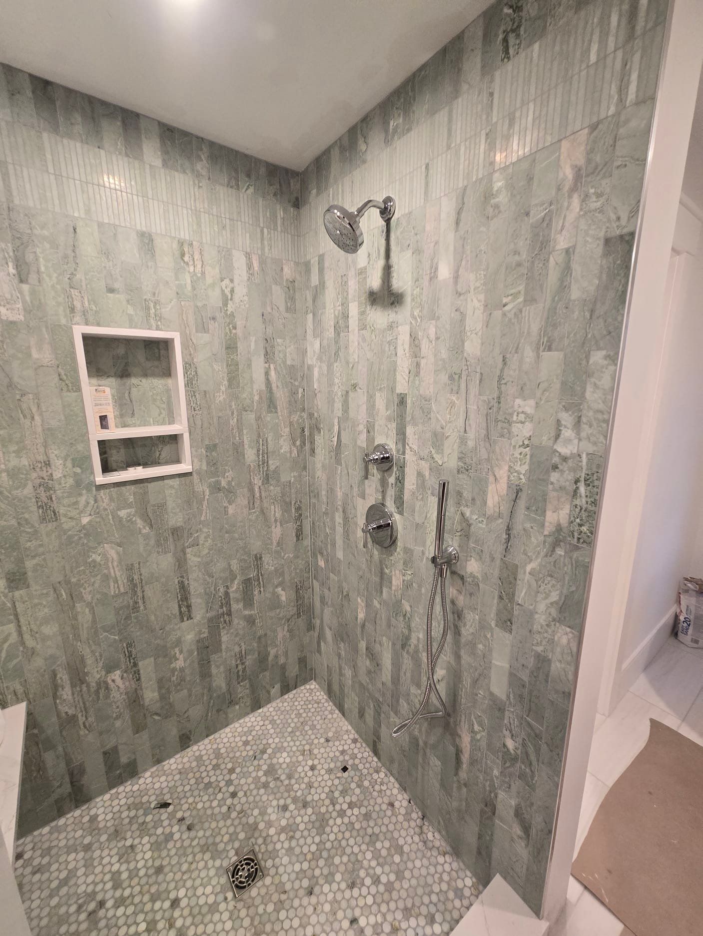 A shower with light green, vertical rectangular tiles on the walls, a white recessed shelf, and a pebbled floor.