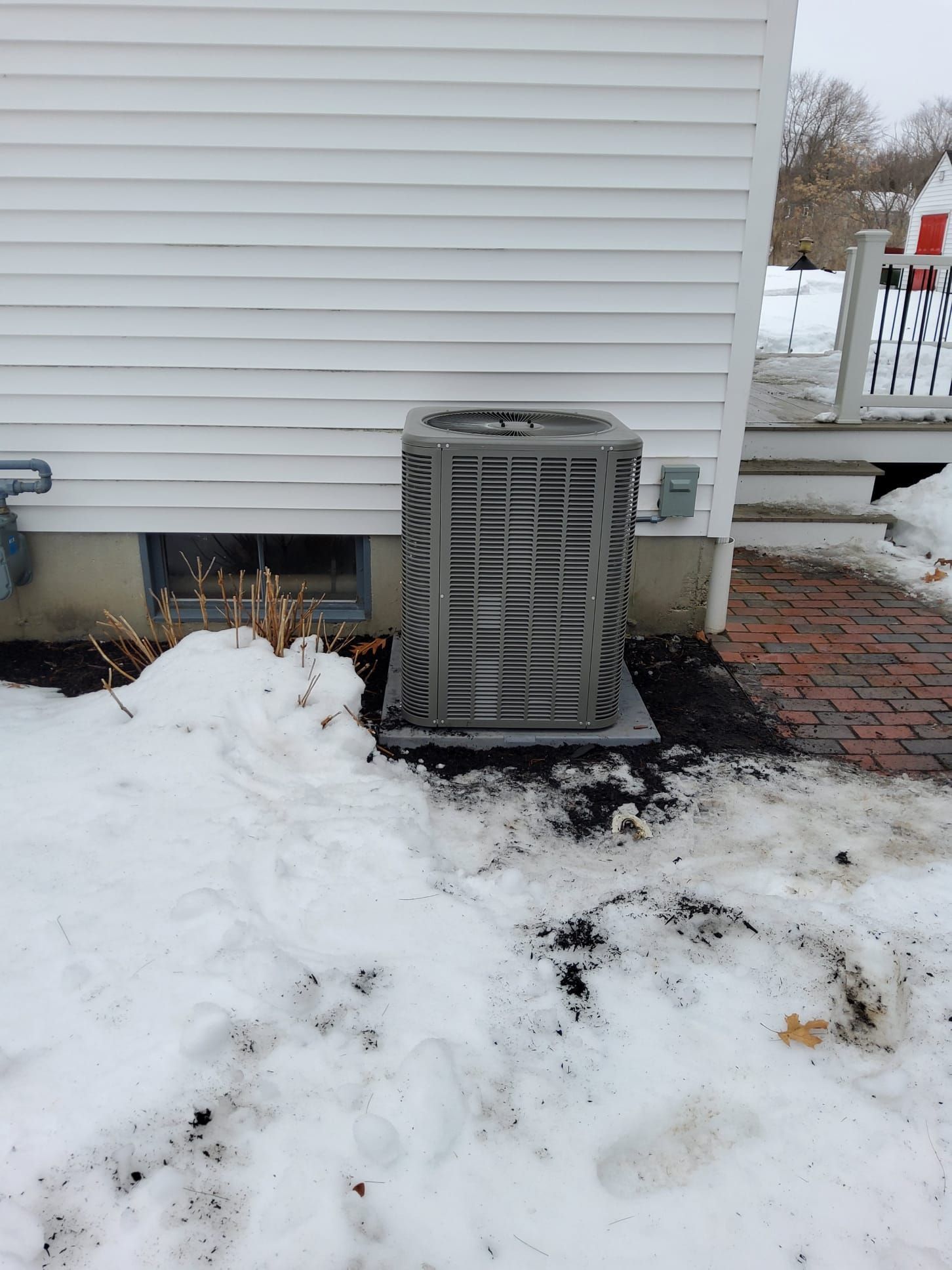 A grey central air conditioning unit sits on a concrete pad against white siding on a snowy winter day.