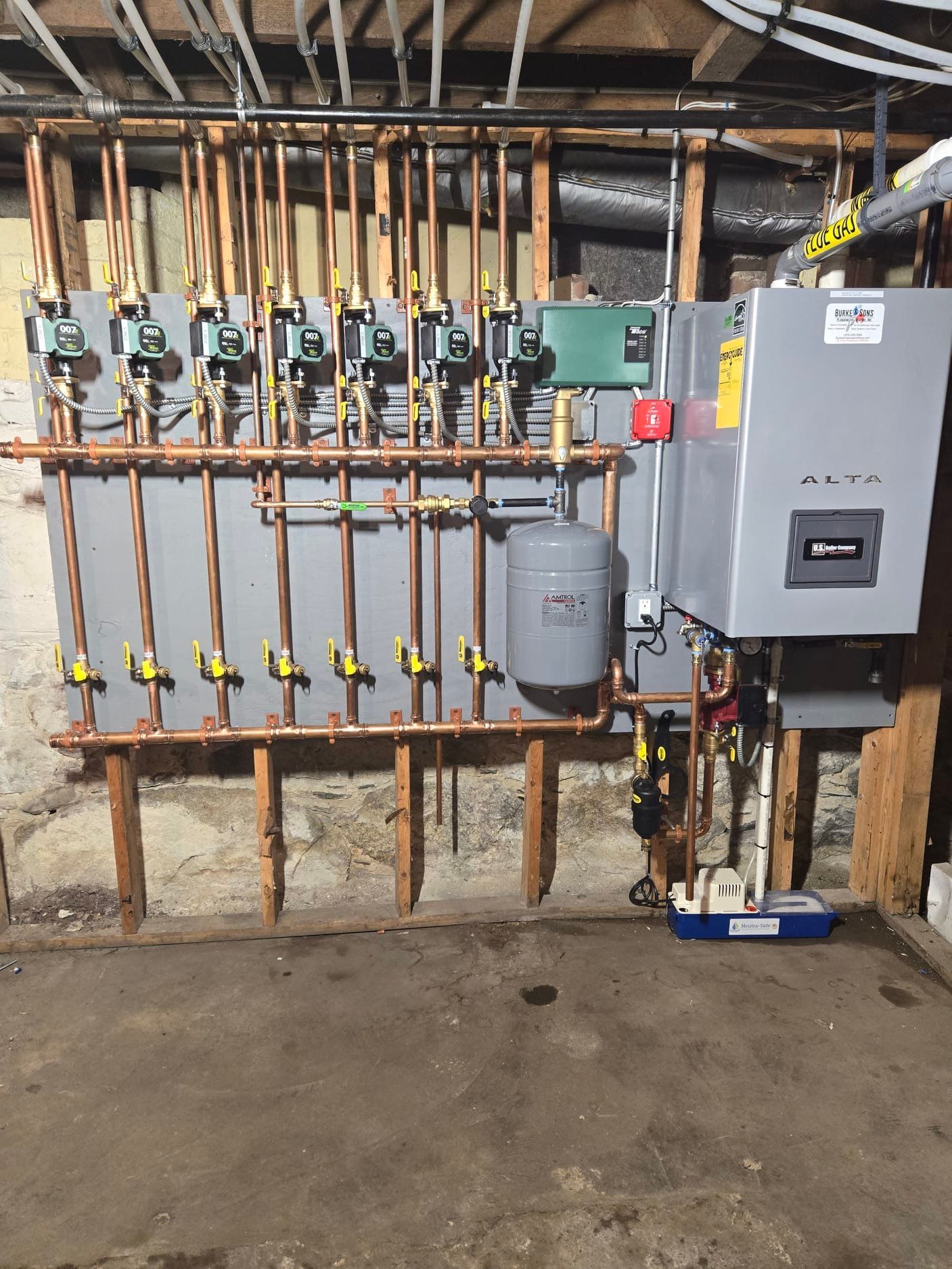 A hydronic heating system with a gray boiler, expansion tank, and a series of copper pipes and pumps on a basement wall.