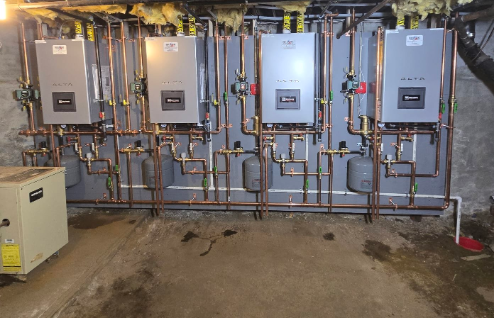 Four wall-mounted boiler units connected by extensive copper piping on a gray panel in a basement.