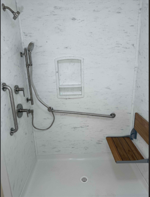 Accessible shower with white walls, a fold-down wooden seat, stainless steel grab bars, and a handheld shower fixture.
