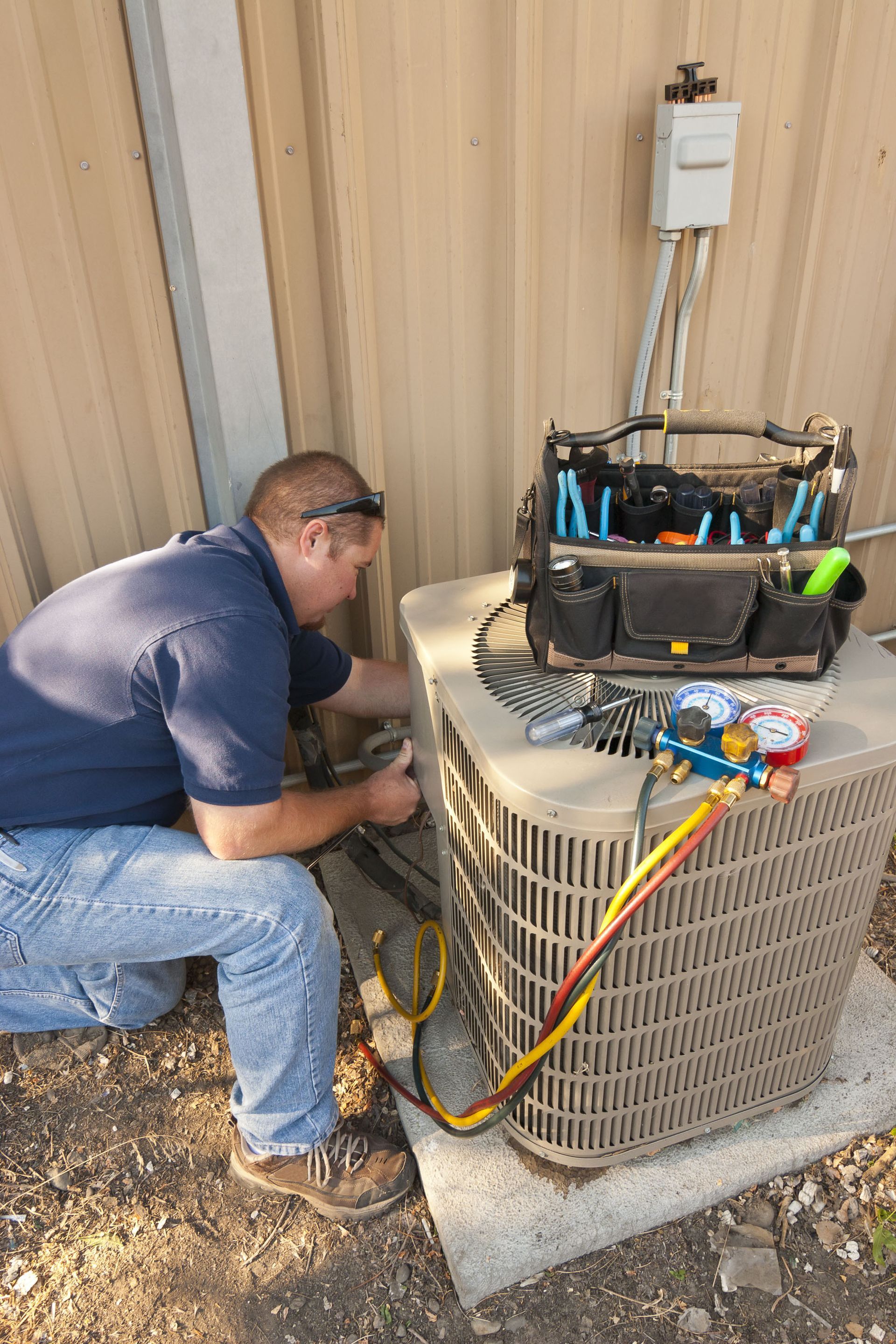 A technician in a blue shirt kneels outdoors to repair an air conditioning unit with tools and gauges nearby.