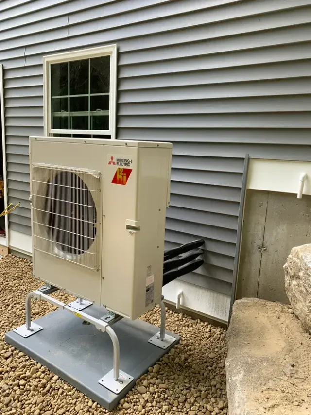 Outdoor mini-split heat pump unit mounted on a raised platform beside a house wall and window