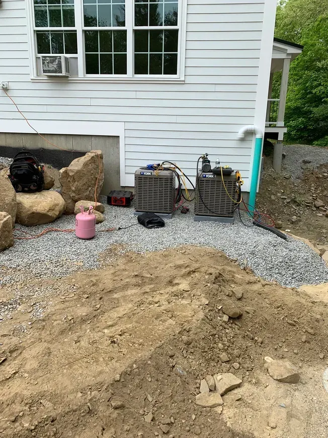Backyard side of a white house with gravel, large rocks, utility units, and a dirt pile under the window