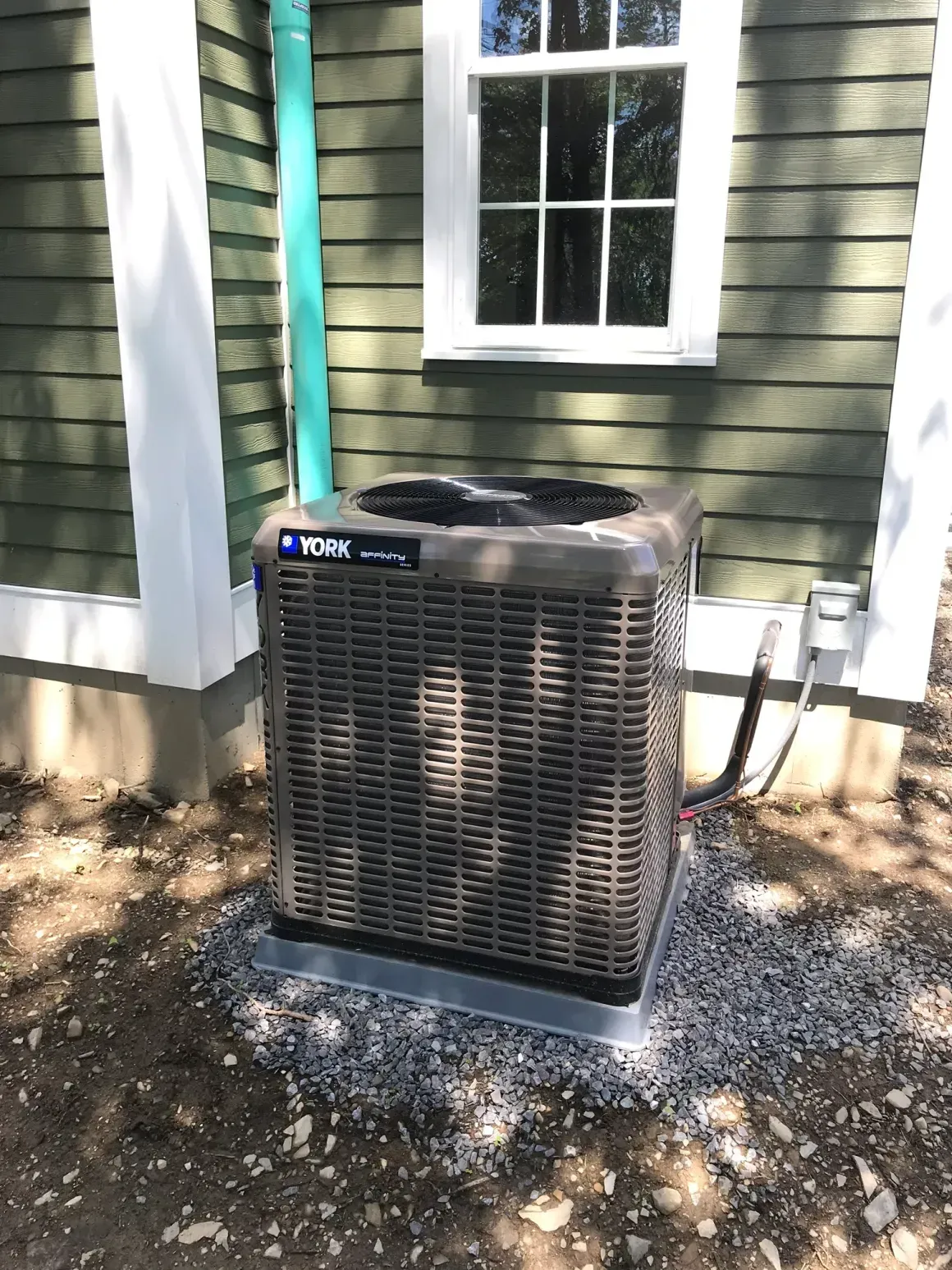 Outdoor air conditioner unit beside a house with green siding and white-trimmed window