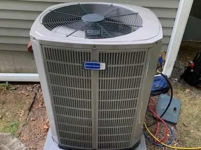 Outdoor air conditioner condenser unit with fan and refrigerant lines on a house exterior.