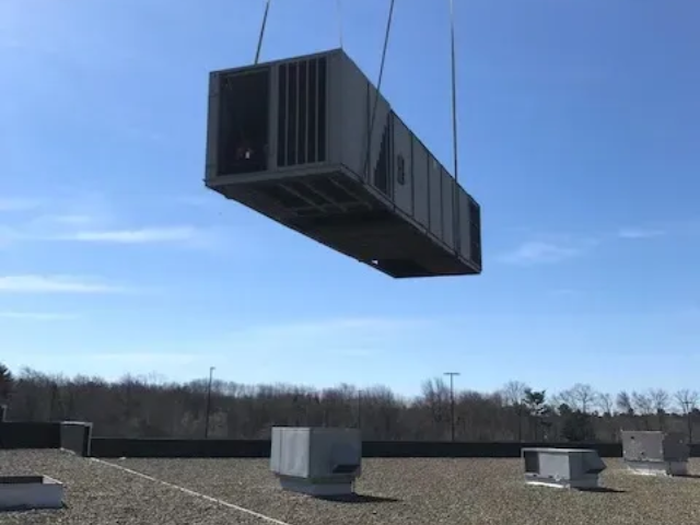 Large black HVAC unit lifted by a crane over a flat rooftop under a blue sky