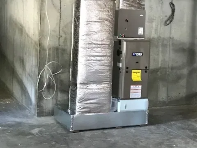 Industrial HVAC unit and ductwork in a gray utility room with concrete walls.