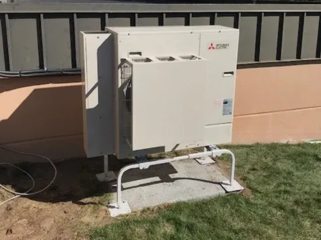Outdoor Mitsubishi air conditioner unit on a concrete pad beside a beige building and grass.