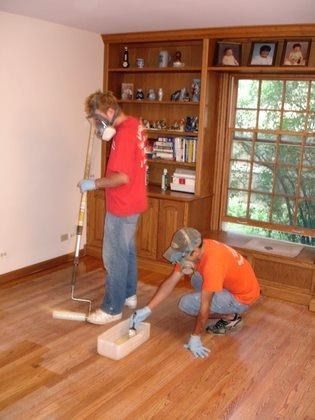 two men are painting a wooden floor in a living room