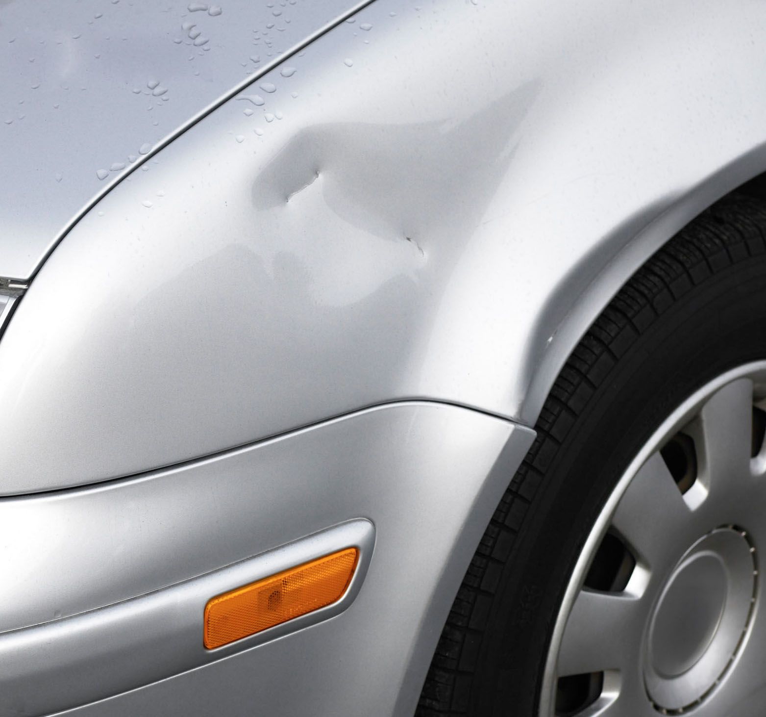 Close-up of a silver car’s damaged front fender with a dent near the wheel and orange side marker.