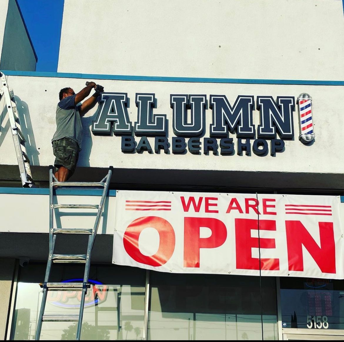 A man on a ladder is working on a sign for alumni barbershop