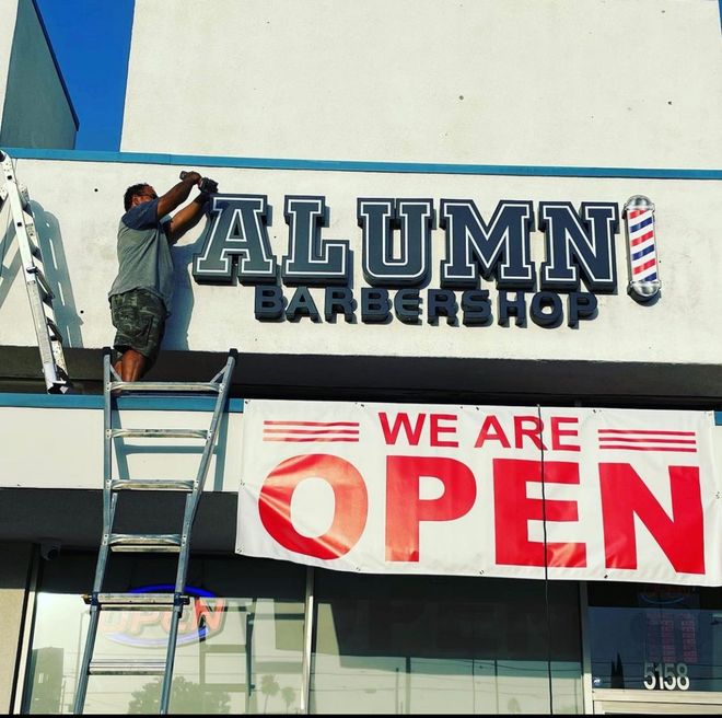 A man on a ladder is working on a sign for alumni barbershop