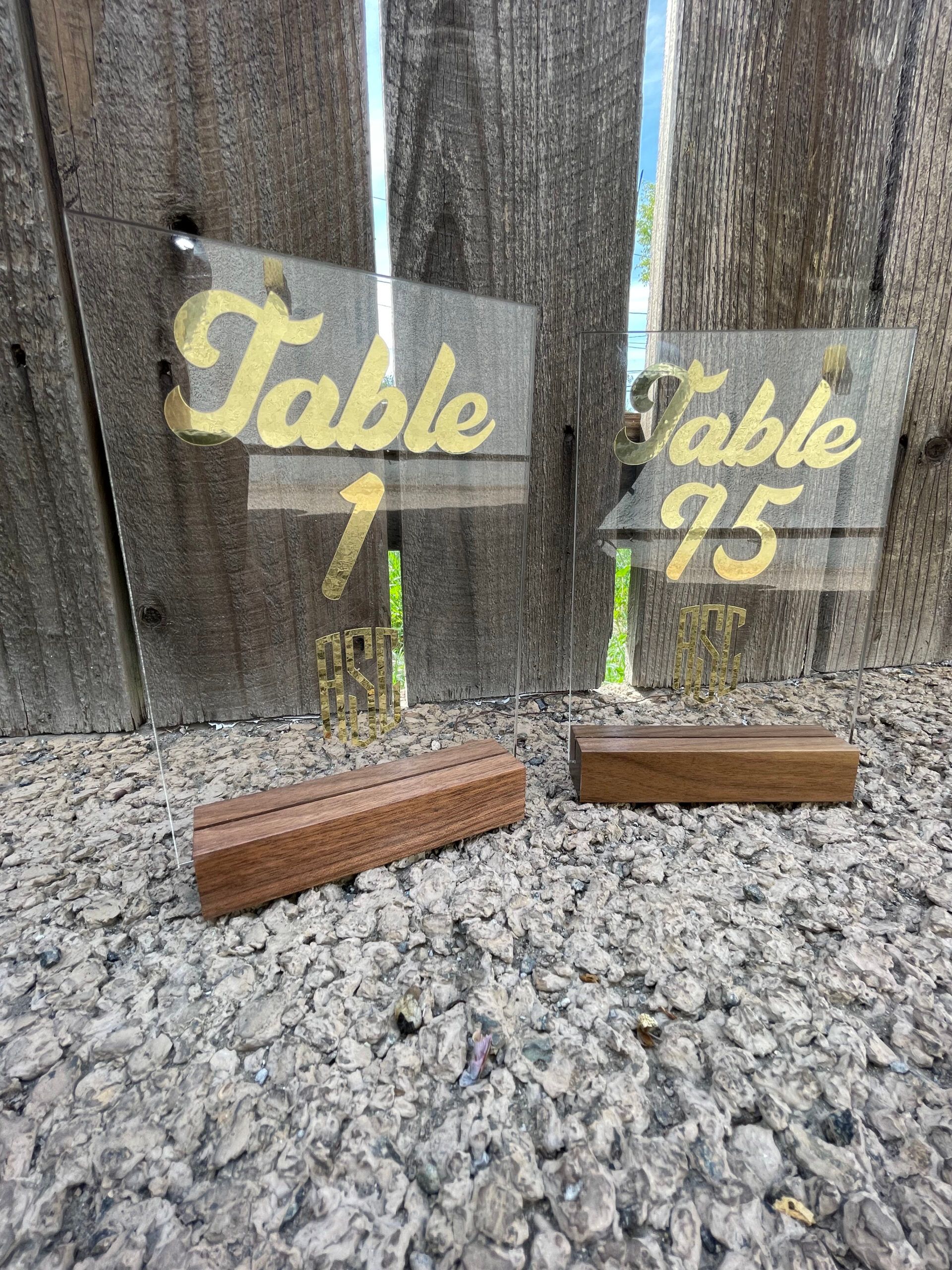Two clear acrylic table numbers with wooden bases are sitting on gravel in front of a wooden fence.