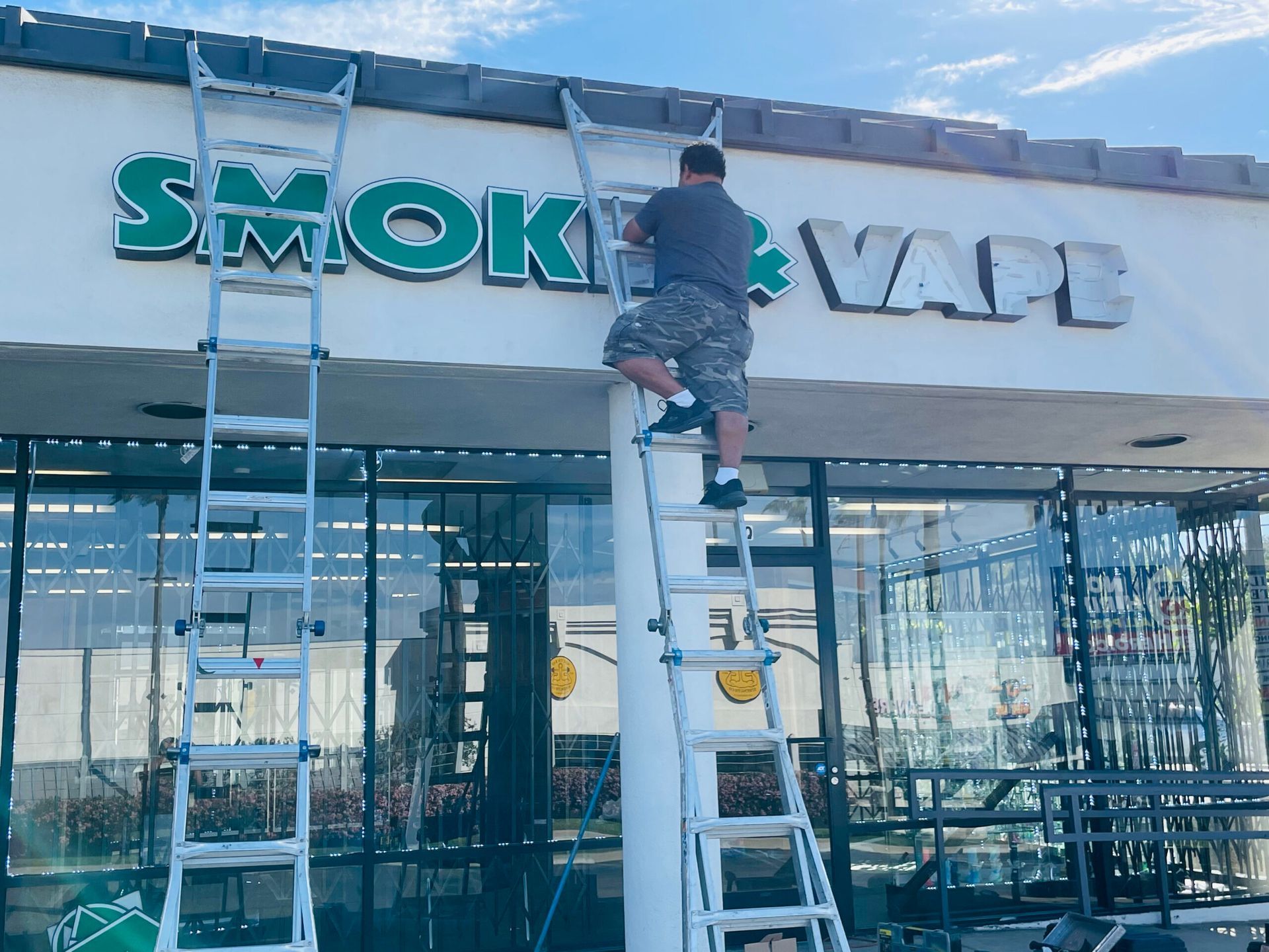 A man on a ladder is working on a sign for smoke vape