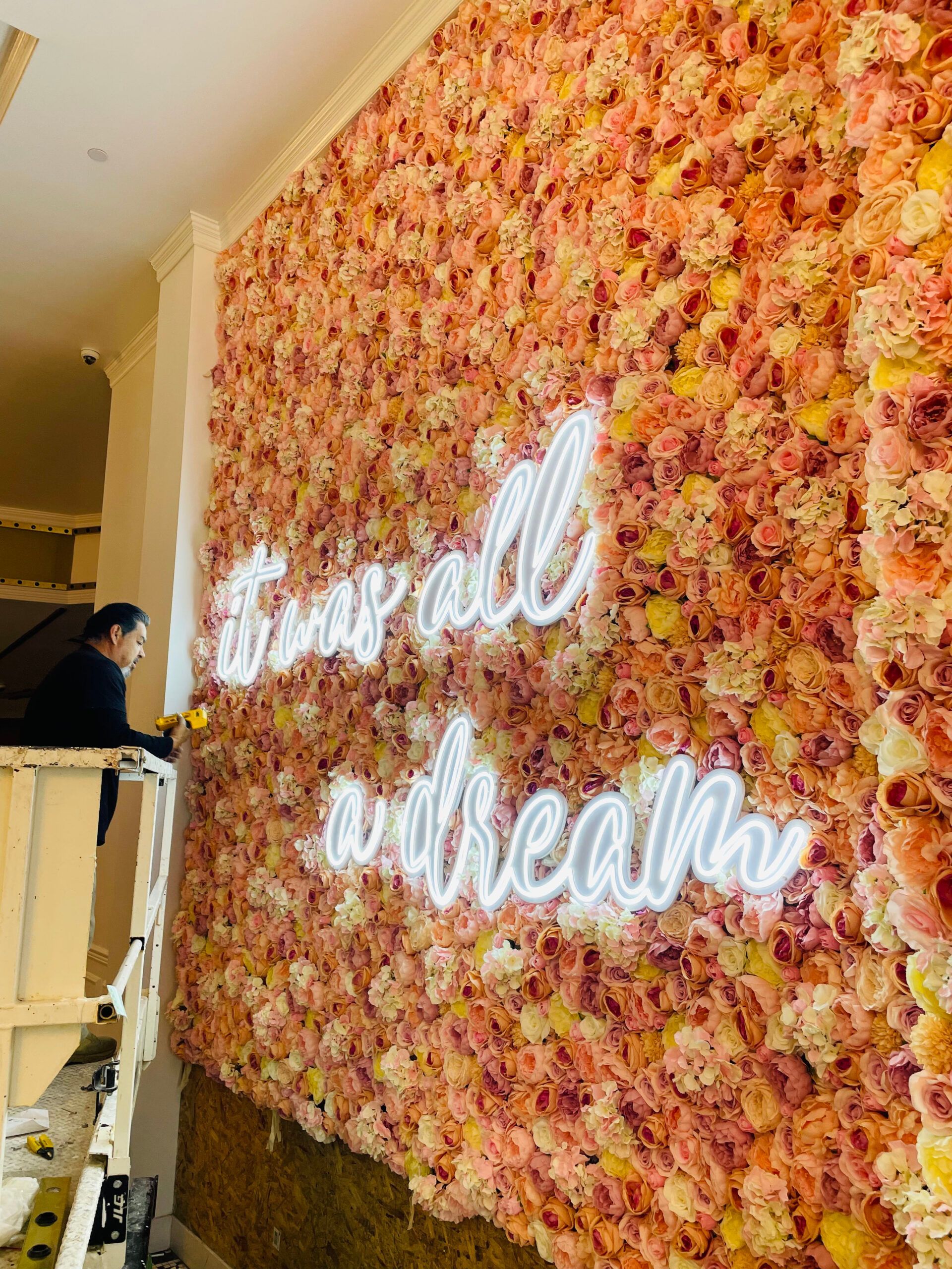 A man is installing a neon sign on a wall of flowers.