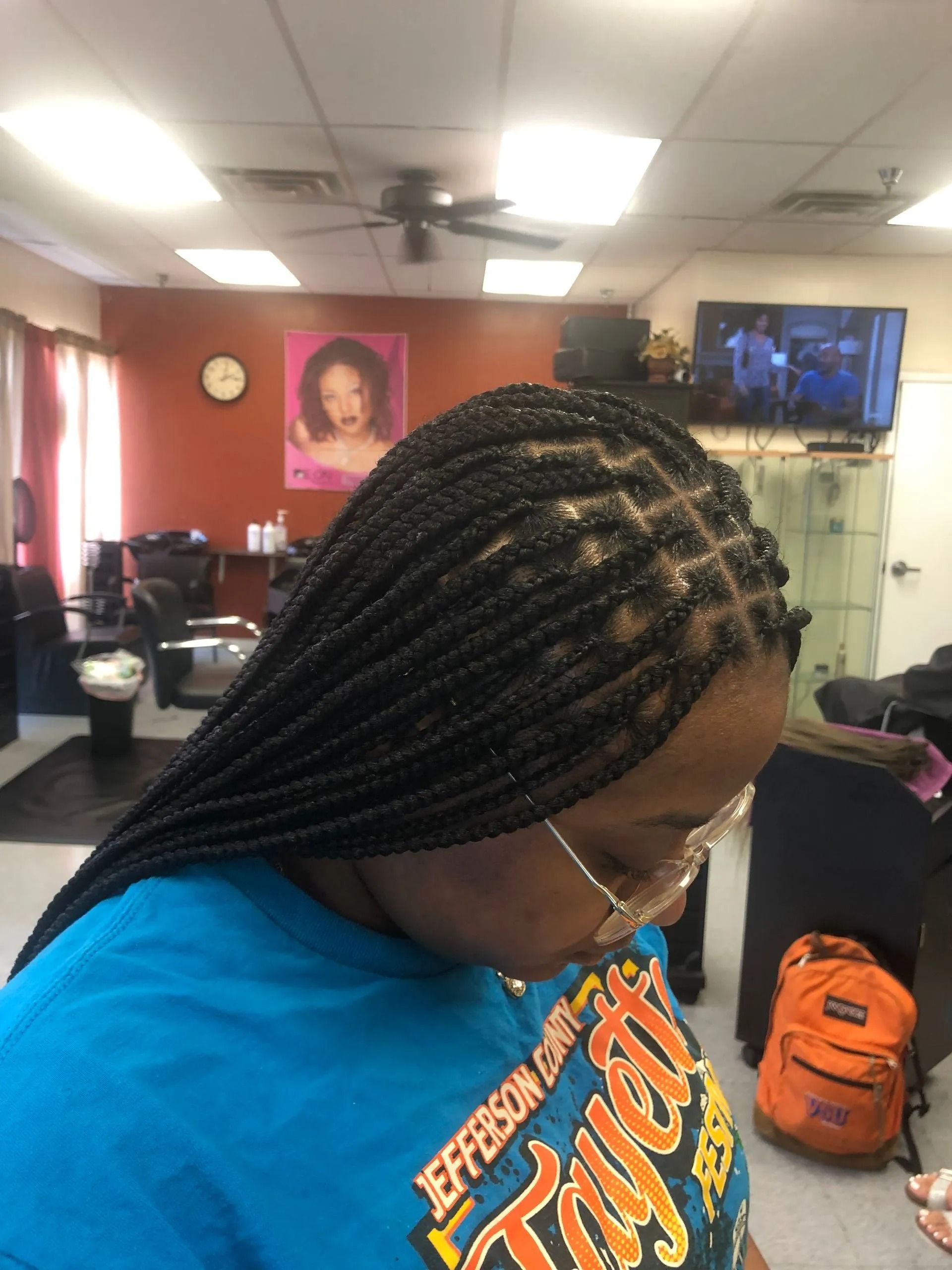 Woman with box braids in a salon, wearing glasses and a blue shirt, orange backpack in background.