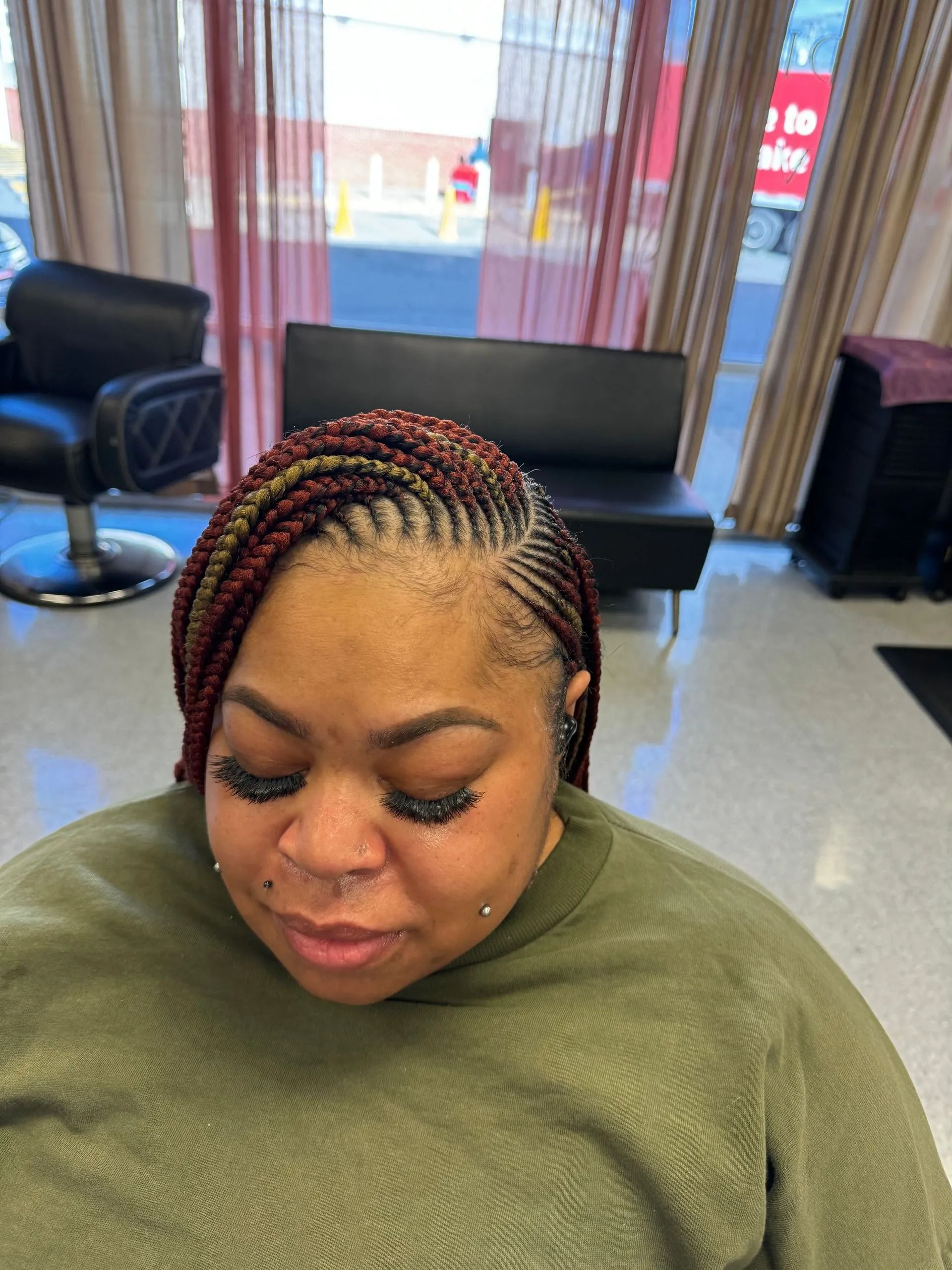Woman with braided hair, wearing green top, in a salon. Braids are red, black and gold.