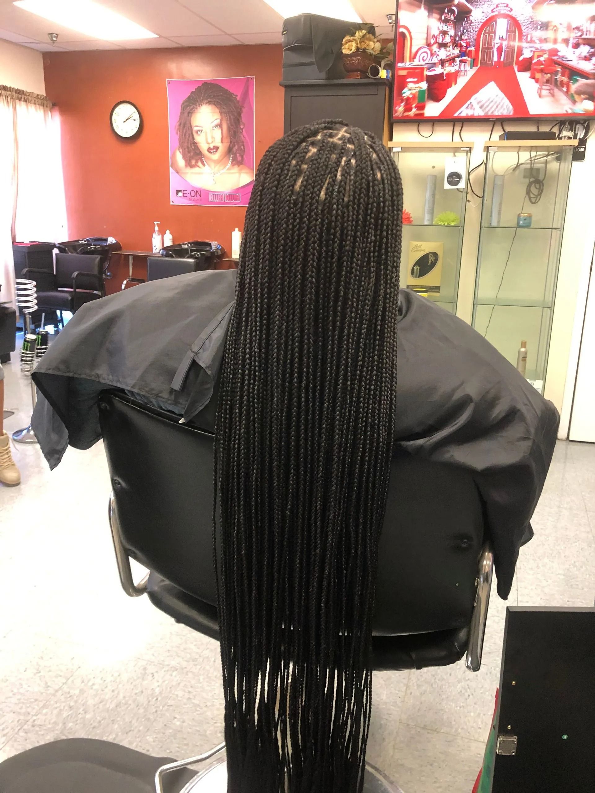 Person in a salon chair with long, black box braids.  A black cape is worn. Interior with decor and a TV.