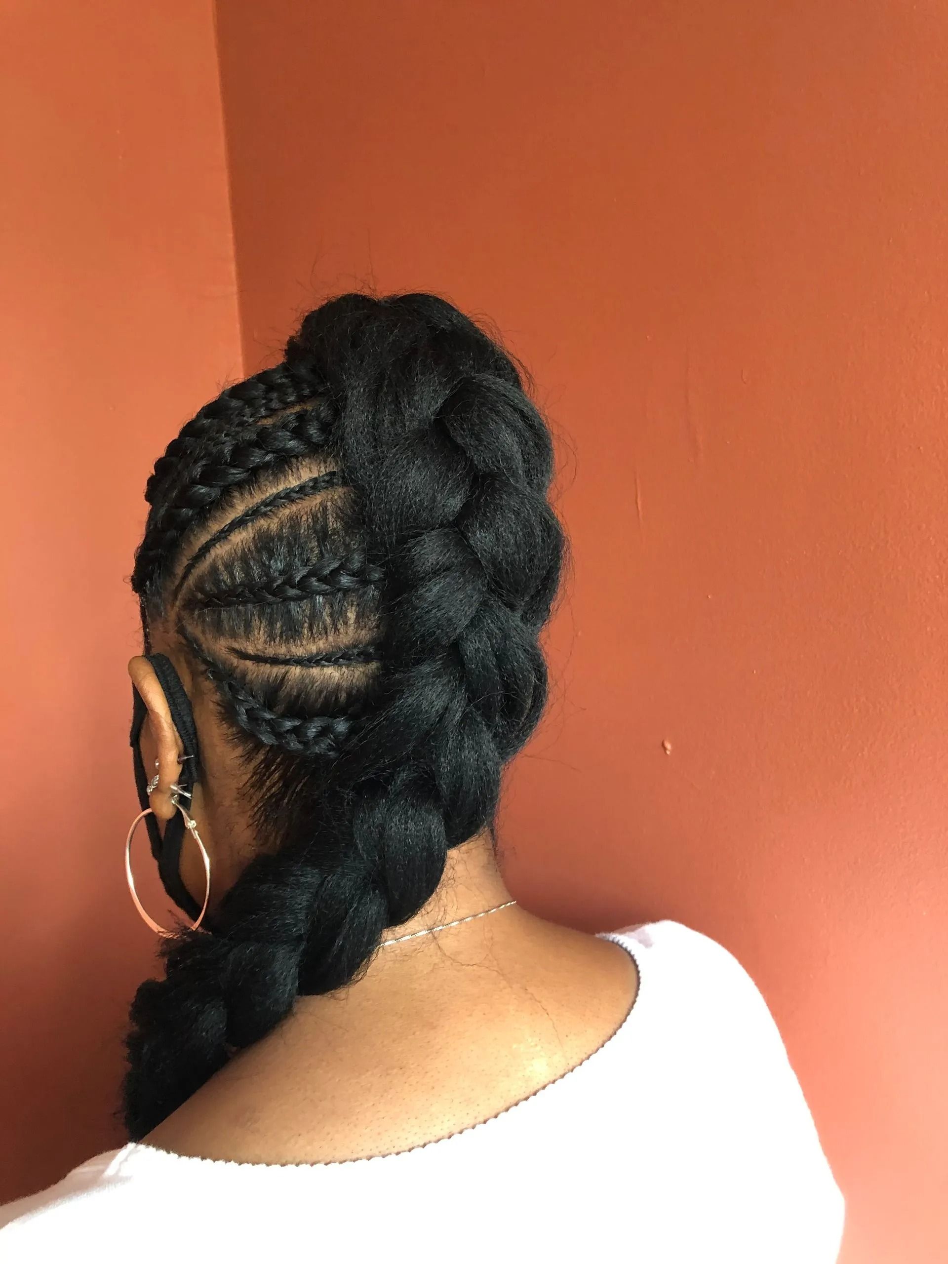Woman with cornrows braided into a side-swept ponytail against an orange wall.