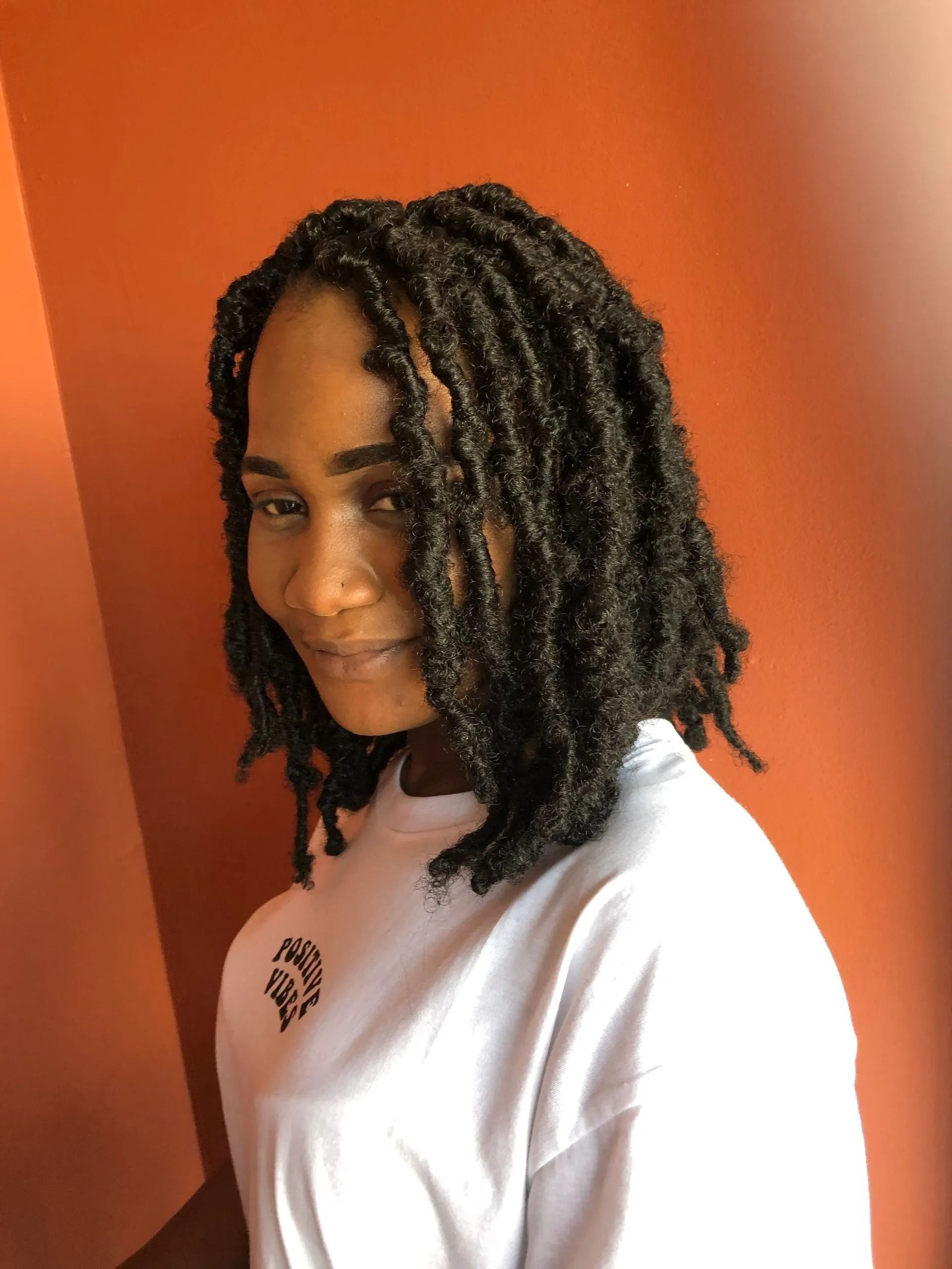 Woman with dark locs smiles, wearing a white tee, against an orange background.