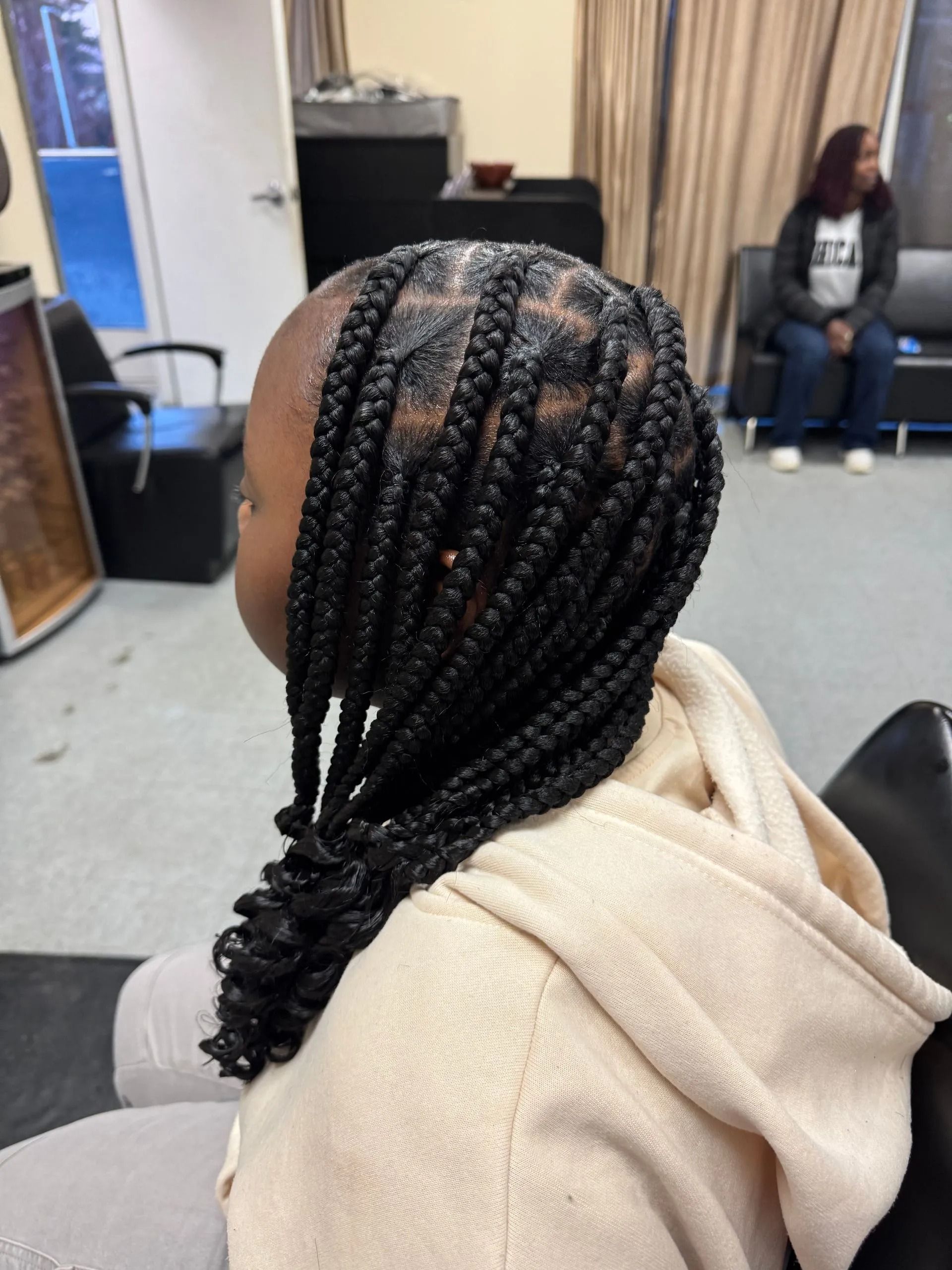 A person with black braided hair. They are sitting in a salon, wearing a tan hoodie.
