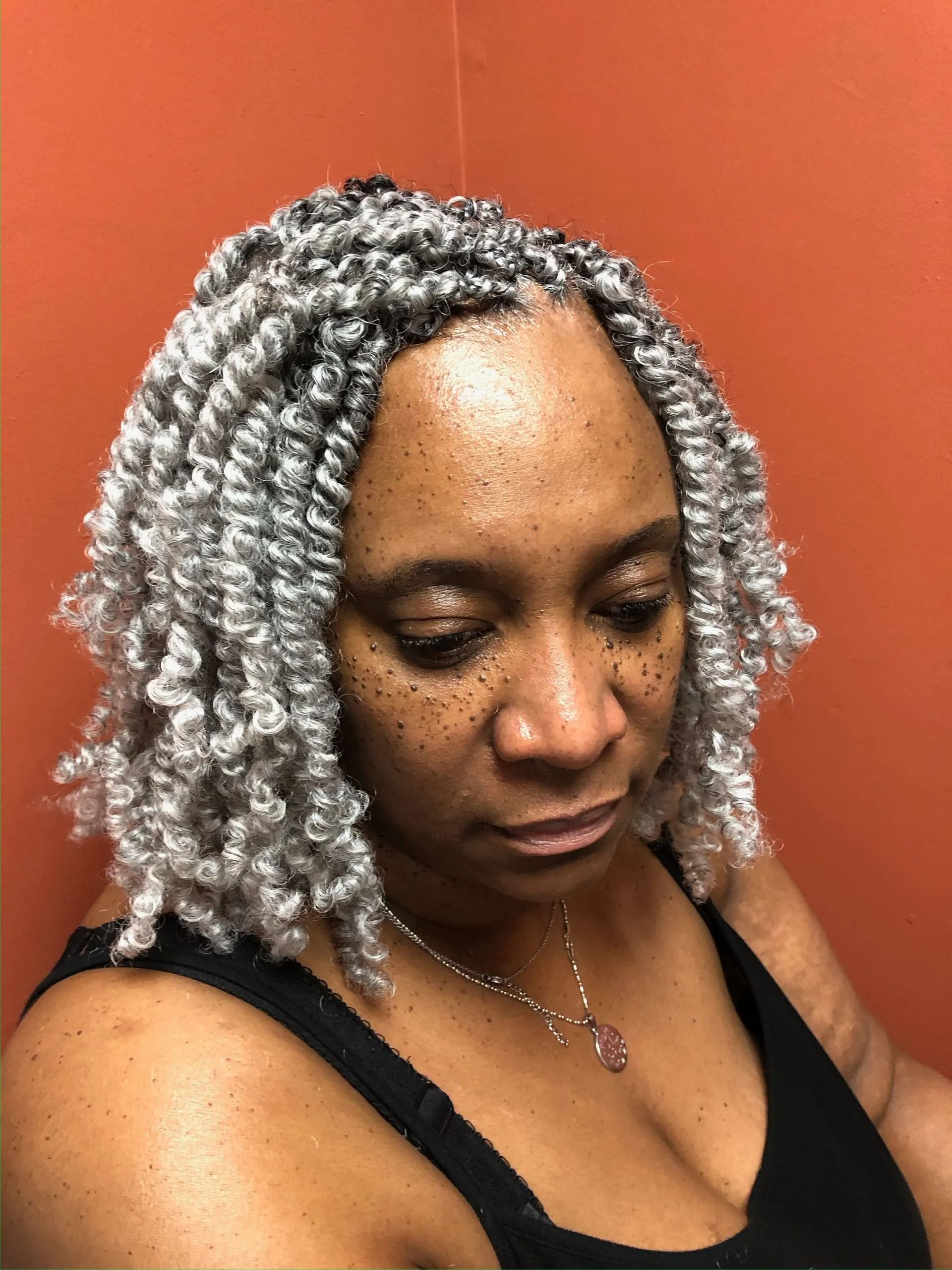 Woman with gray, twisted hair and freckles, wearing a black top against an orange background.