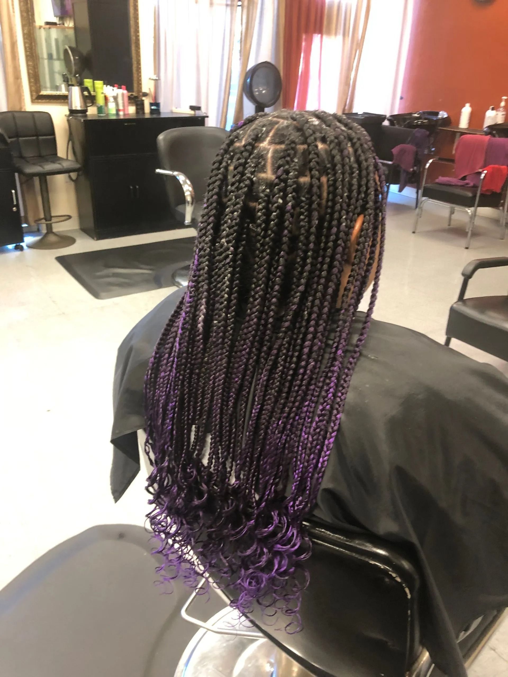 Woman with long, black box braids that transition into purple curls, seated in a salon chair.