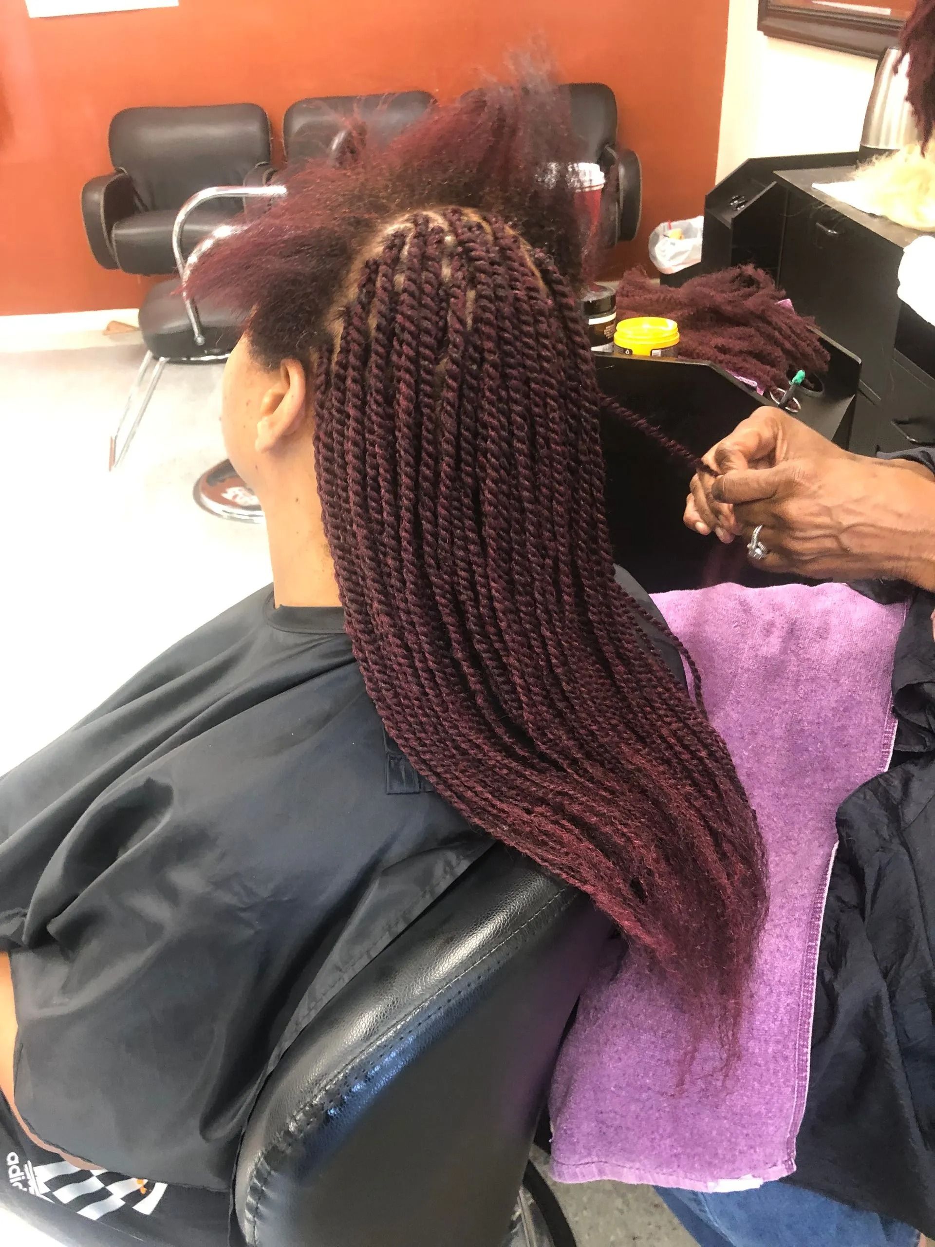 Woman getting burgundy braids in a salon. Braider's hands are visible, hair is draped over a purple towel.