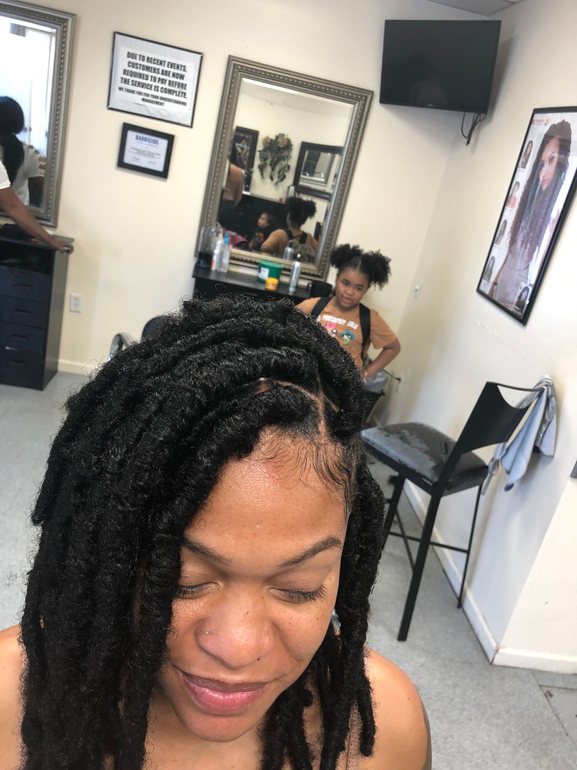 Woman with long, black faux locs at a hair salon. A girl and mirror are in the background.
