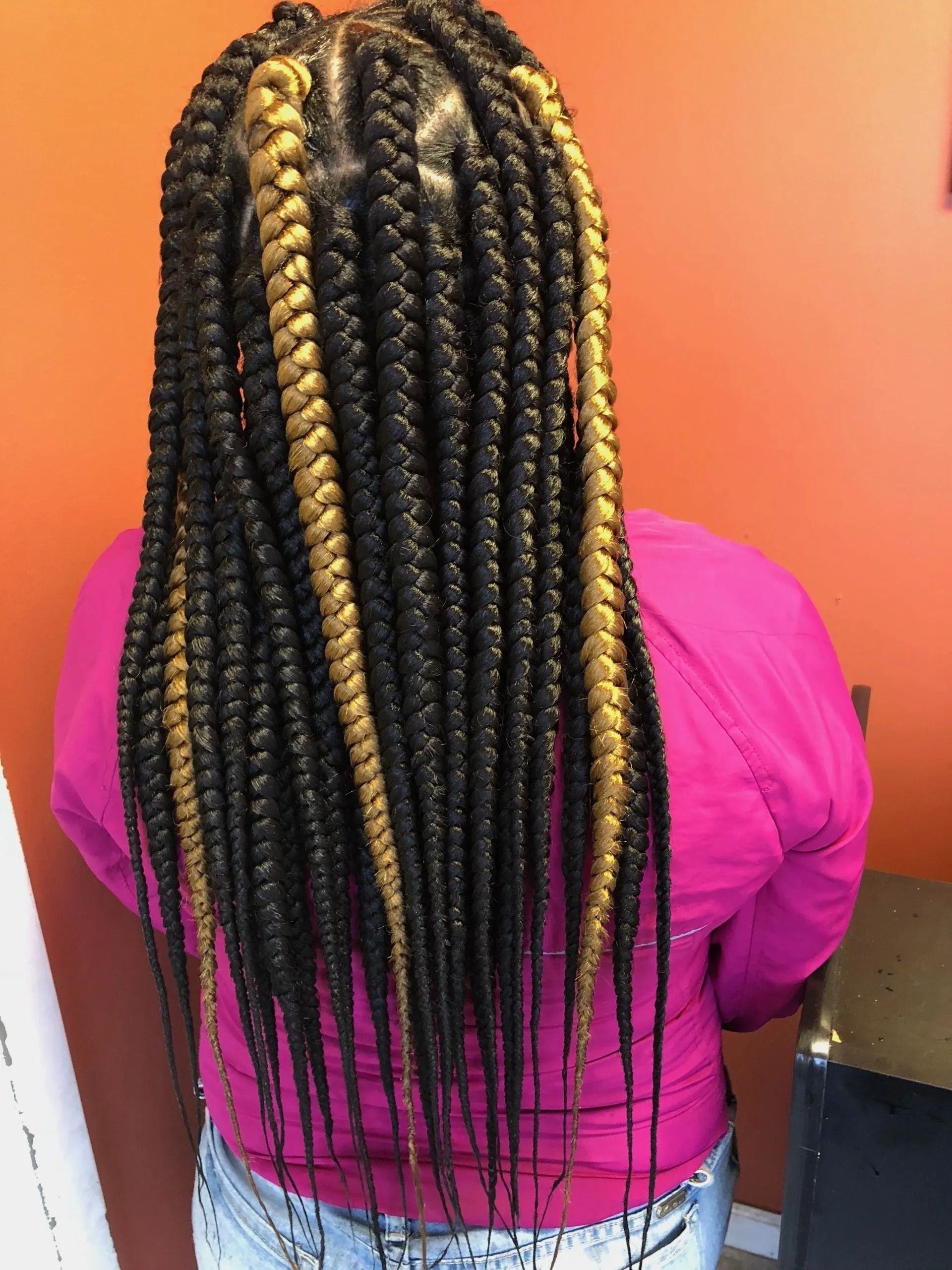 Woman with long black and gold box braids, wearing a pink top, facing an orange wall.