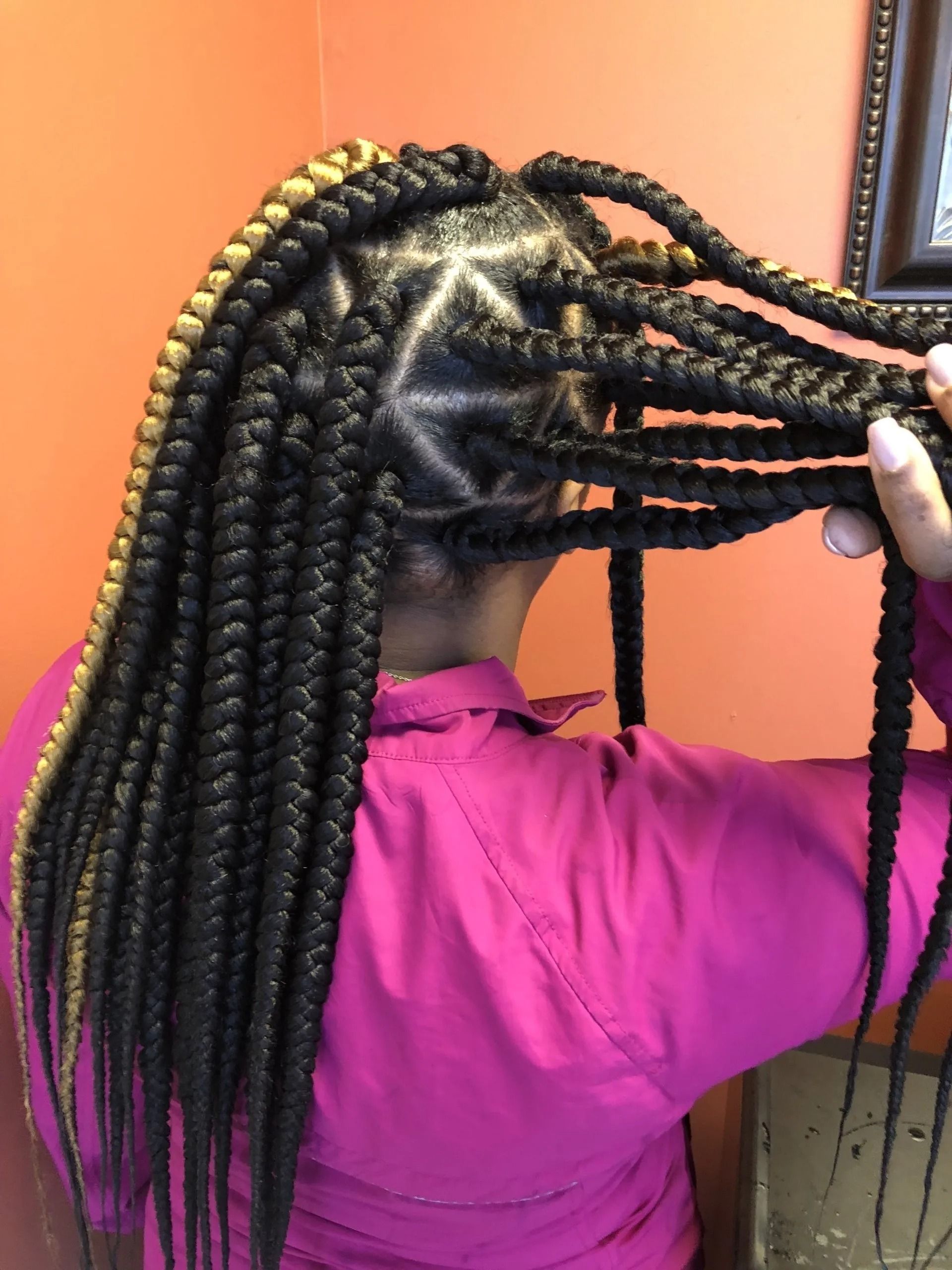 Woman with long, black and blonde box braids, styled with geometric pattern on the side. Wearing a pink top.