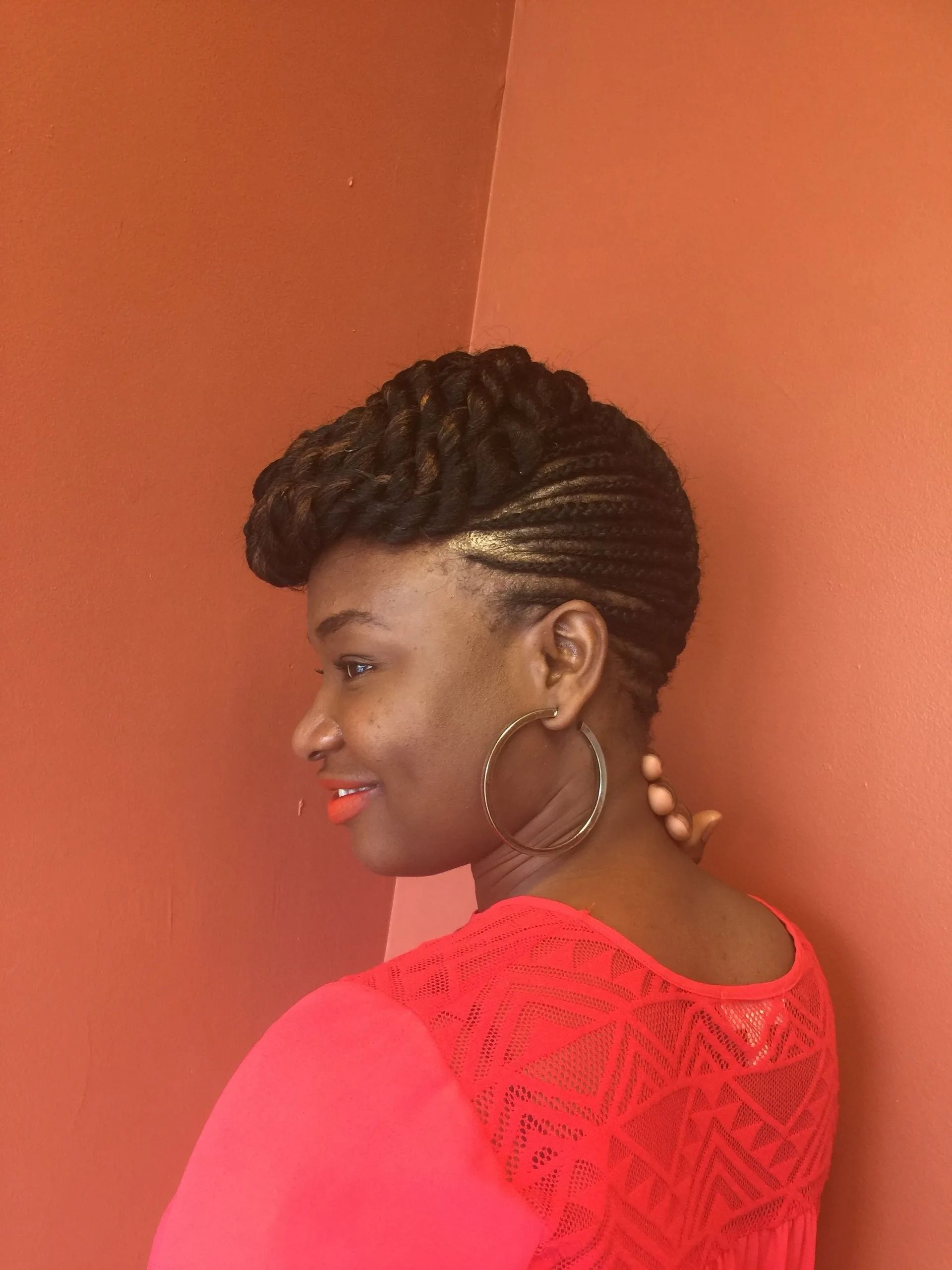 Woman with braided updo smiling, wearing red shirt and hoop earrings. Leaning against orange wall.