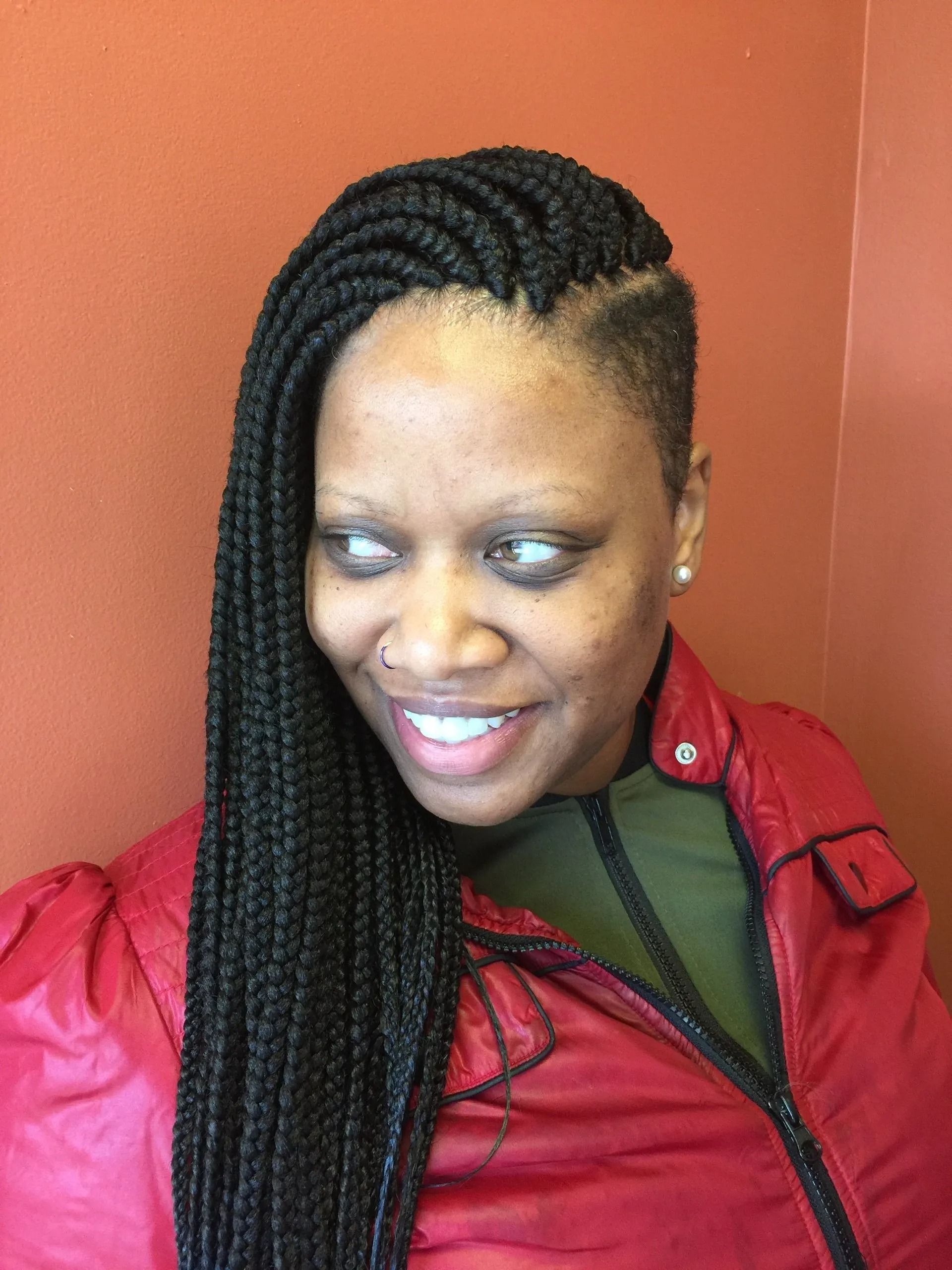 Woman with long black braids and shaved side, wearing a red jacket, smiling.