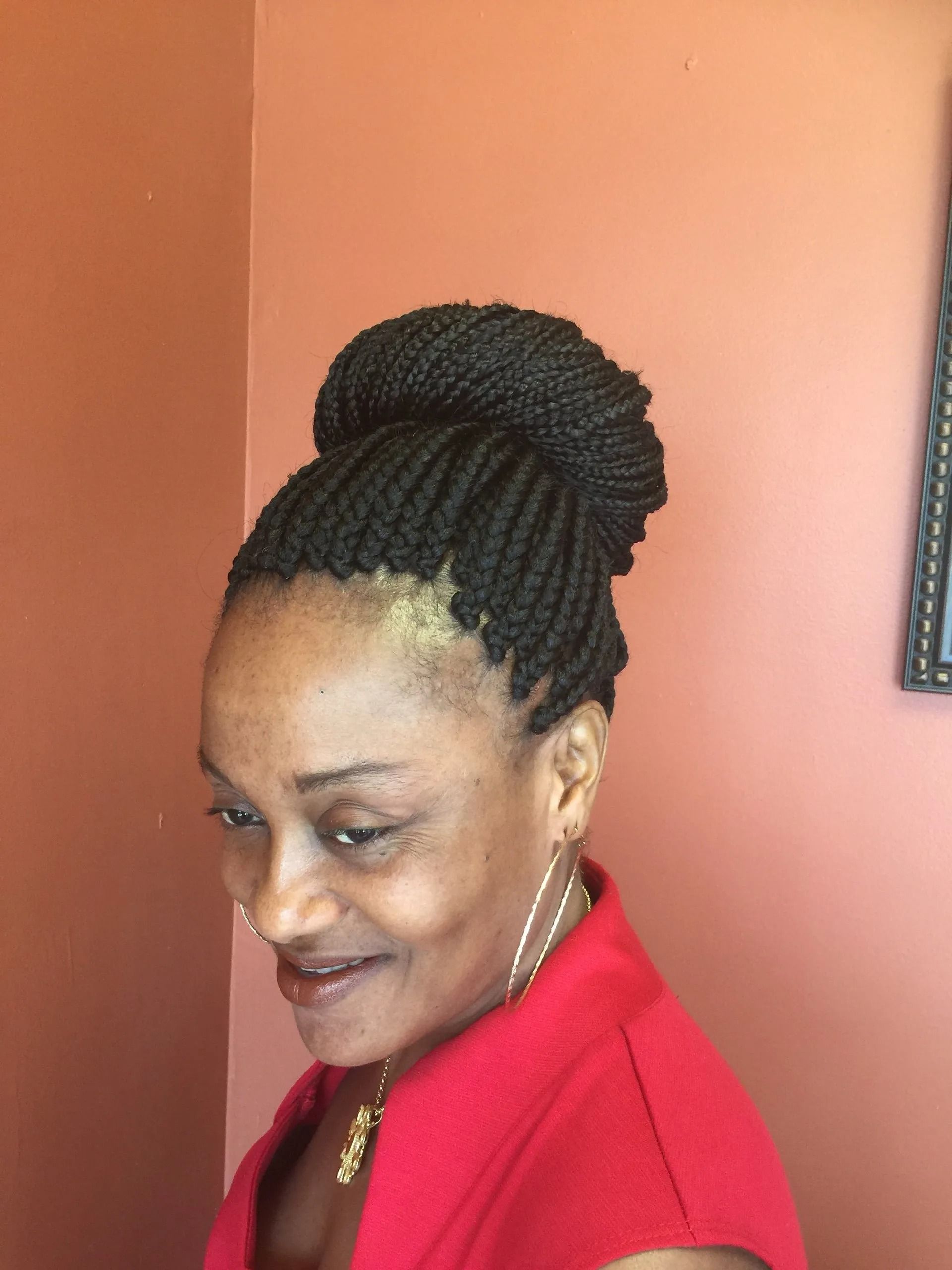 Woman with dark braided hair up in a bun, wearing a red top, against a peach wall.