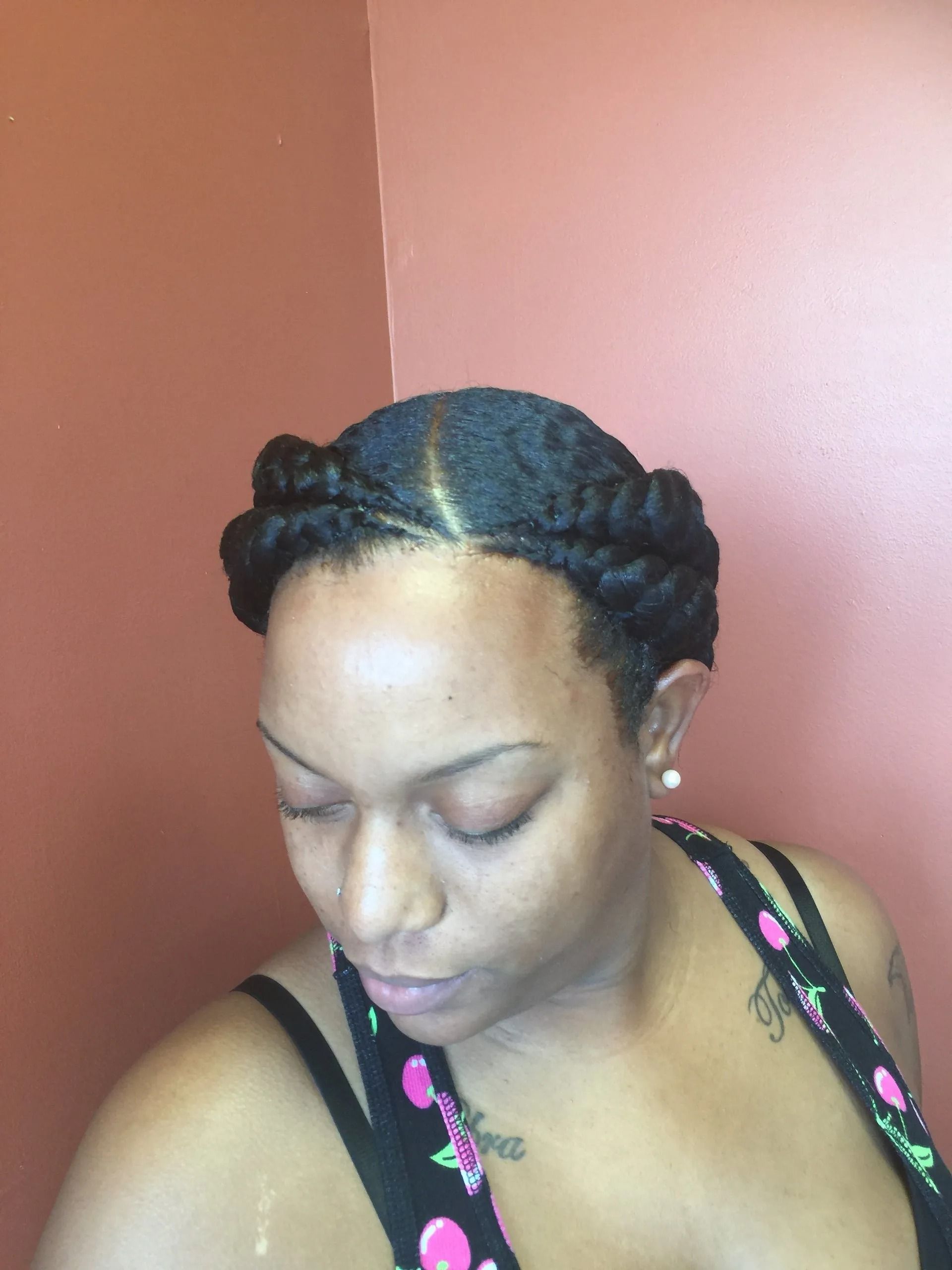 Woman with natural hair in a braided crown style, looking down. Pink wall in the background.