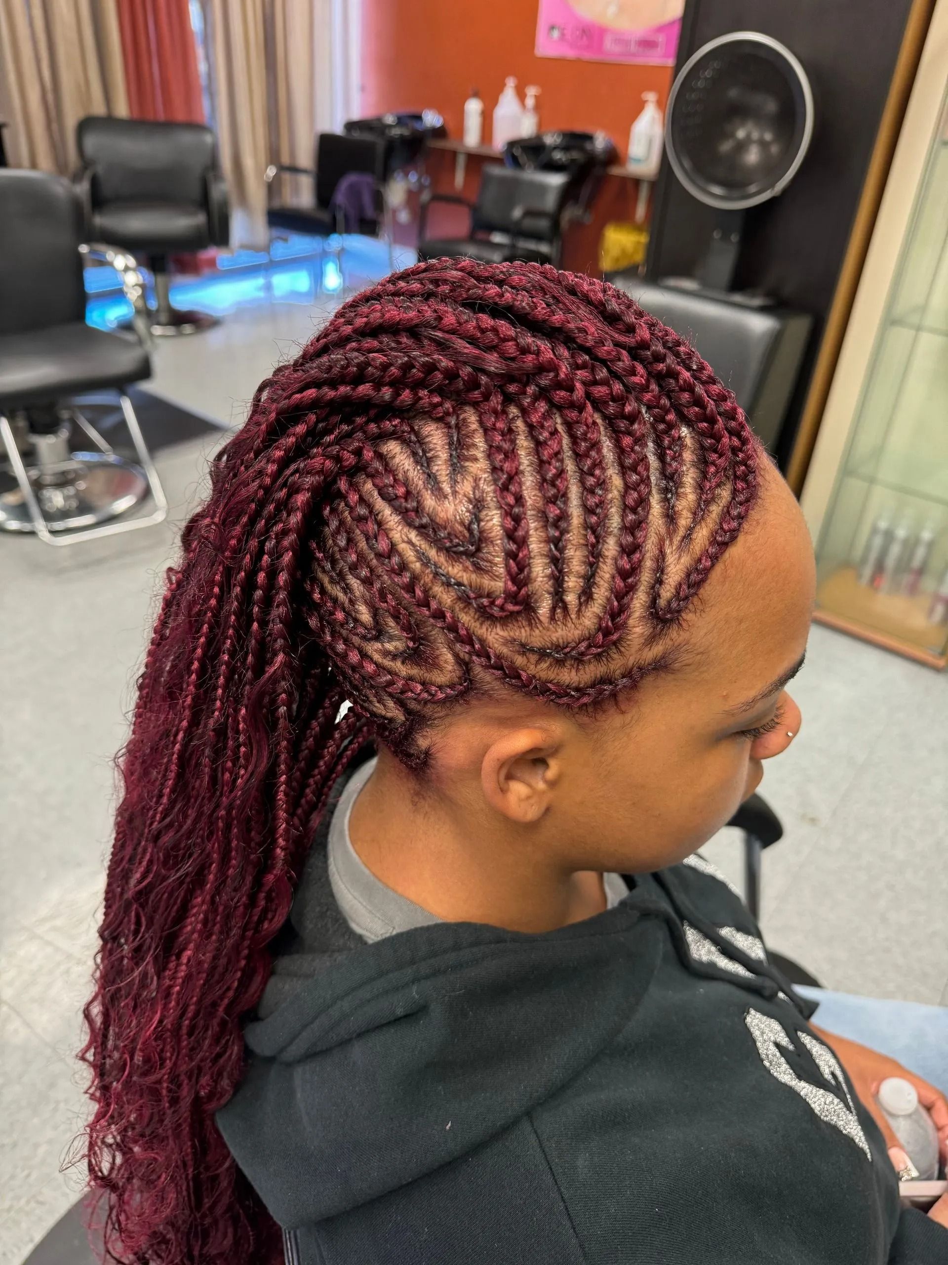 Woman with long burgundy braids, cornrows with a patterned design on the side. Indoors.