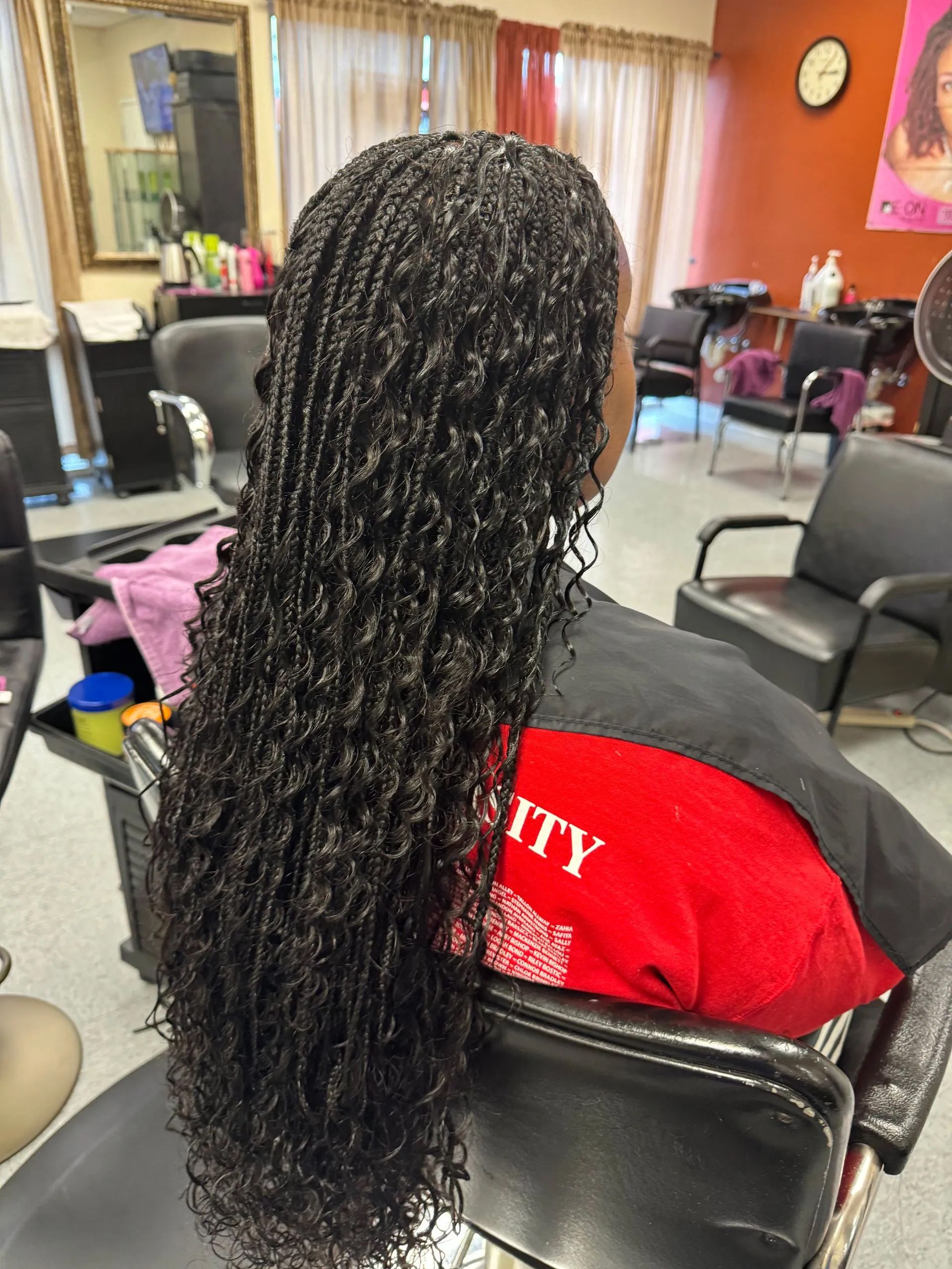 Woman with long, curly micro braids sits in a salon chair. She wears a red and black cape.