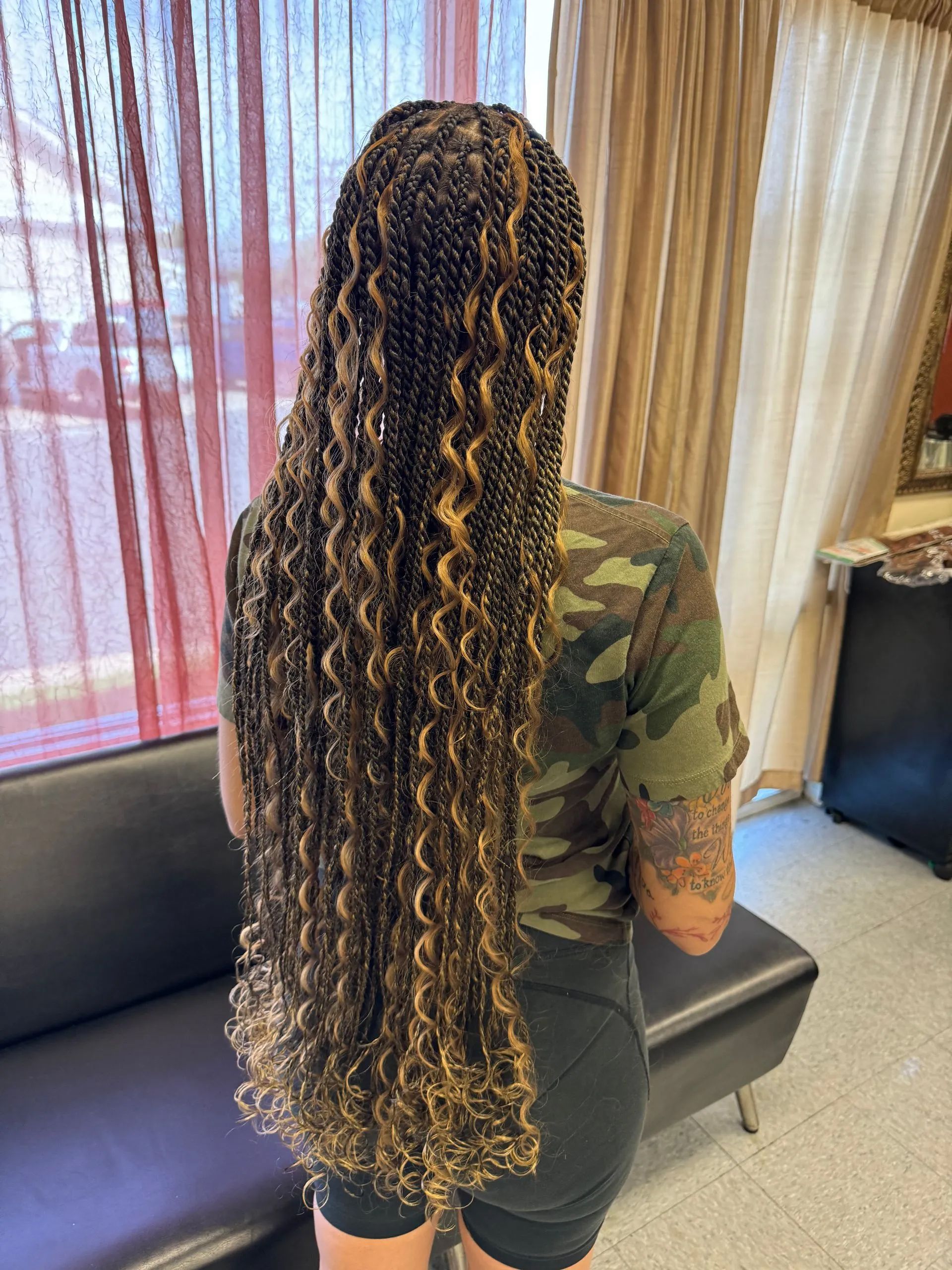 Woman with long, curly, golden-brown braids stands facing away from camera in a salon.