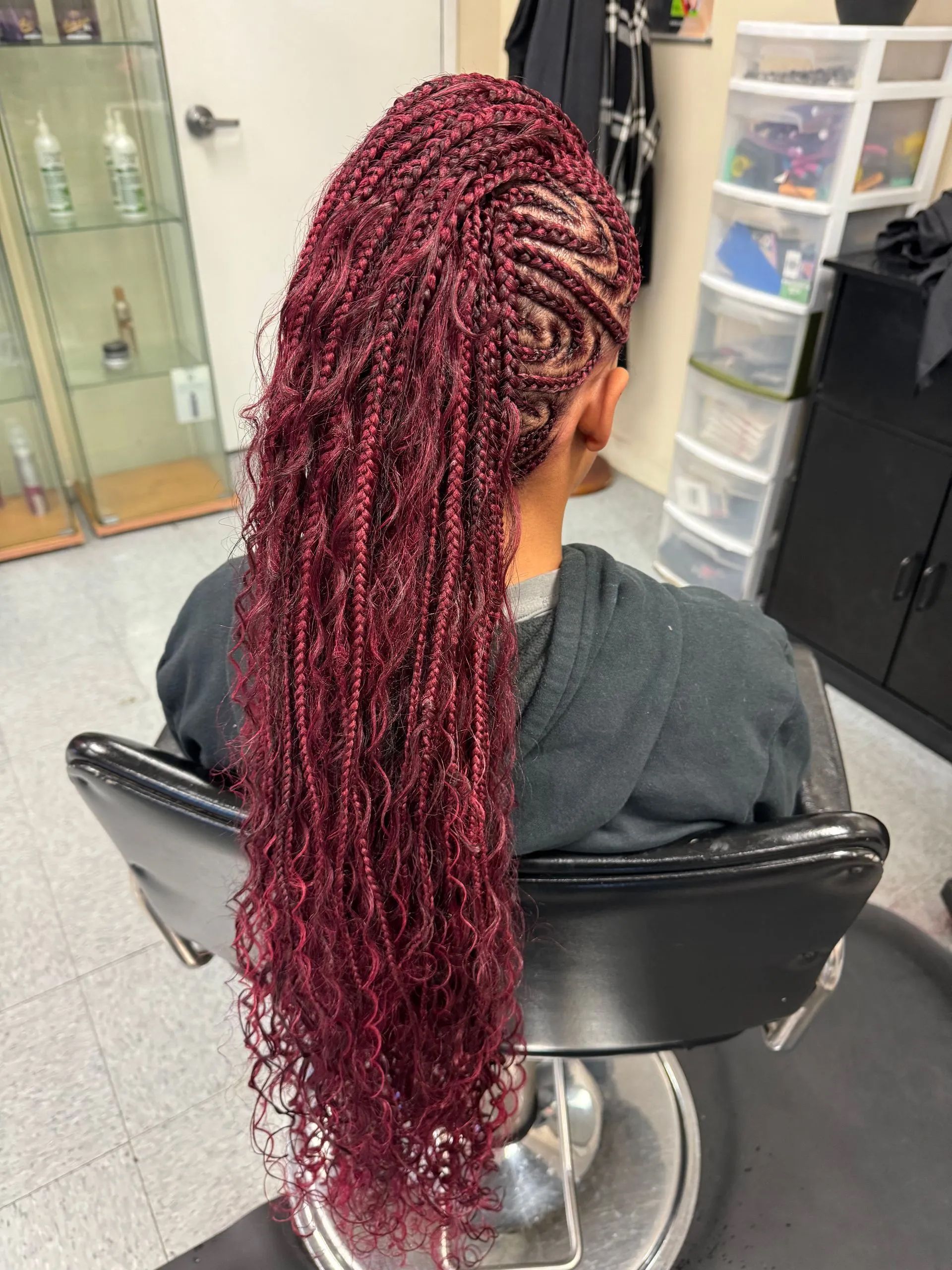 Woman with long, burgundy micro braids, partially braided on one side. Sitting in a salon chair.