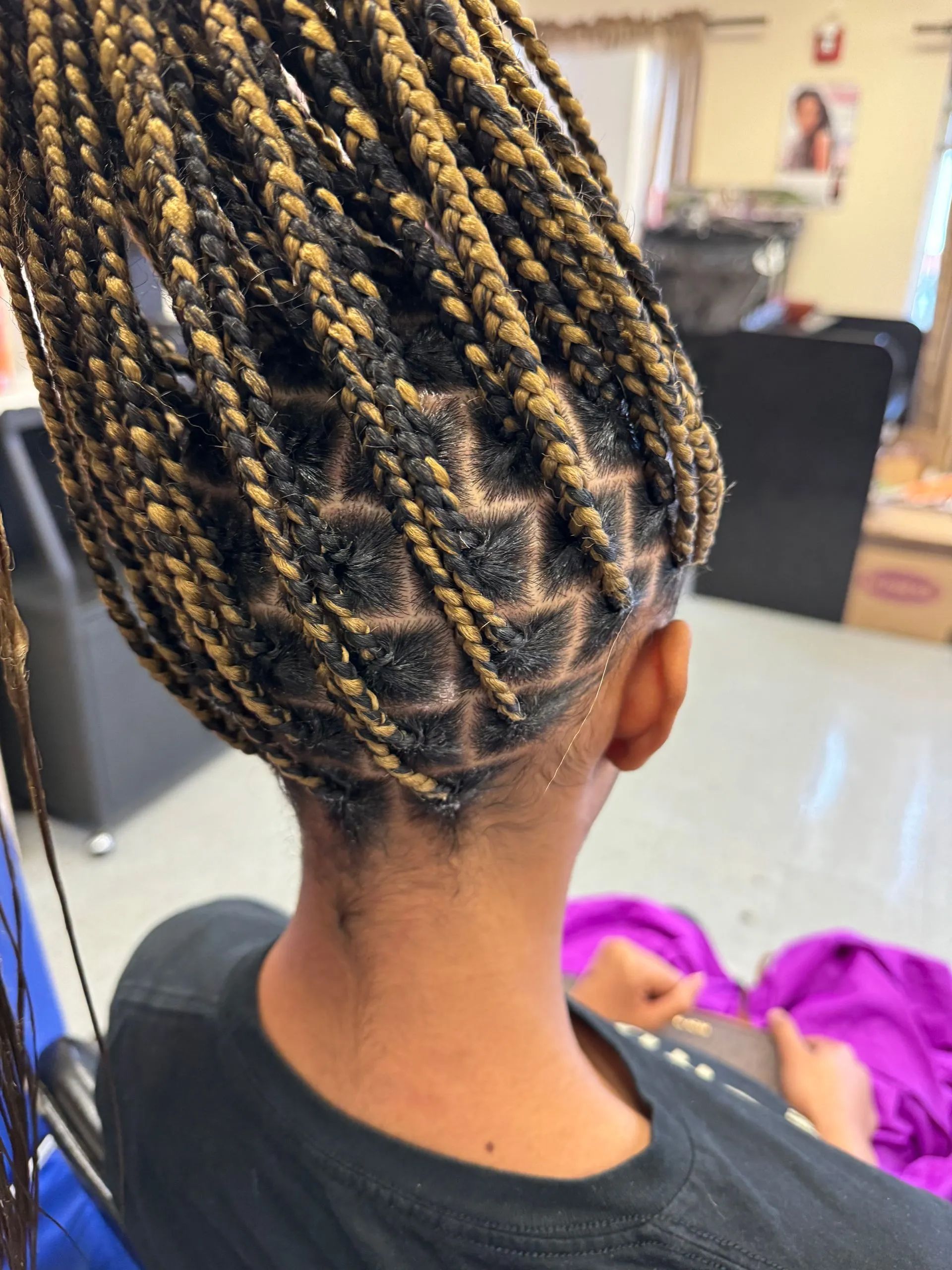 Person with box braids featuring a diamond pattern, with black and blonde hair, sitting in a salon chair.
