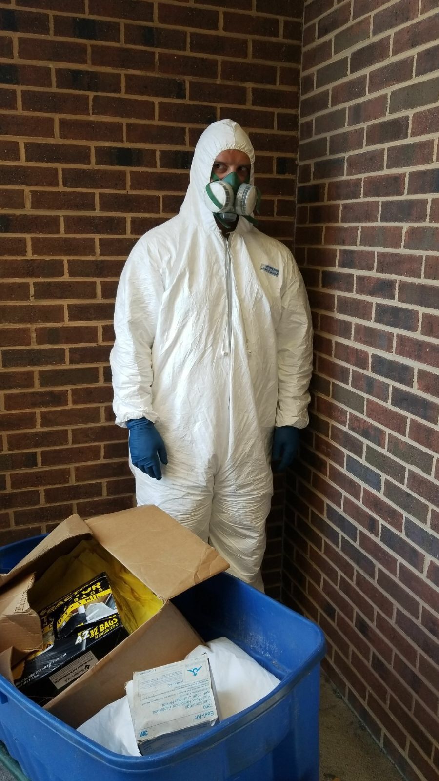 A man in a protective suit is standing next to a blue trash can.