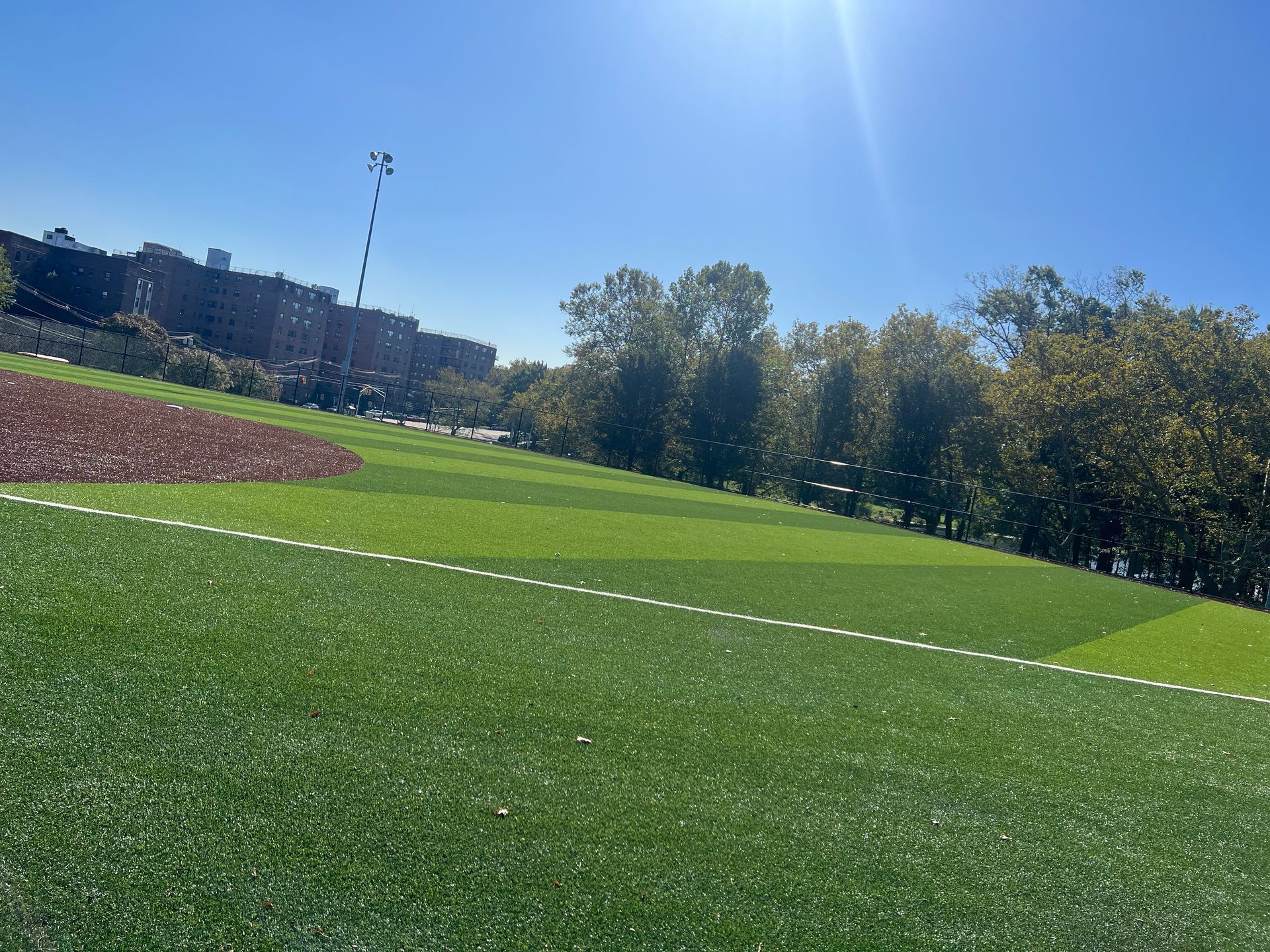 A sunny outdoor scene featuring a green artificial turf field with a red gravel area, lined with trees and buildings.