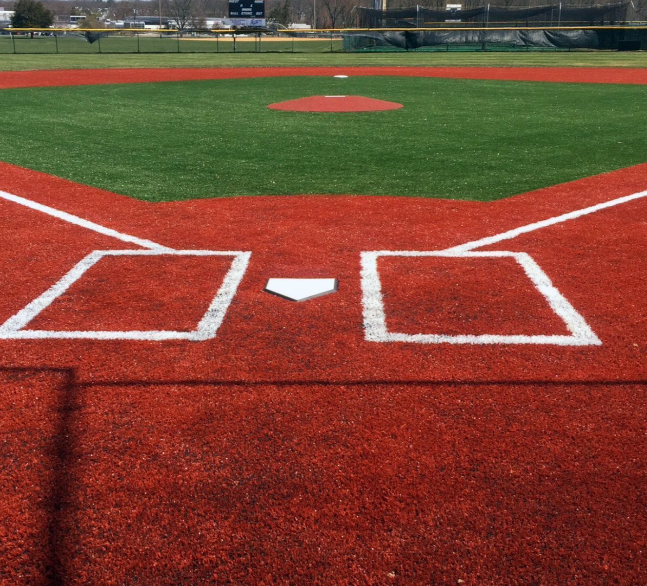 A close-up view of home plate, batter's boxes, and the red infield dirt of a baseball field with green turf in the back.