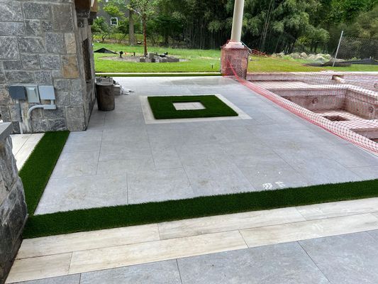 Outdoor patio with grey stone pavers, strips of green artificial grass, and a central square grass inlay near a pool area.