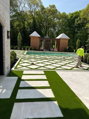 A patio featuring white stone pavers set in a diamond pattern with turf inlays, leading to a pool and two brick structures.