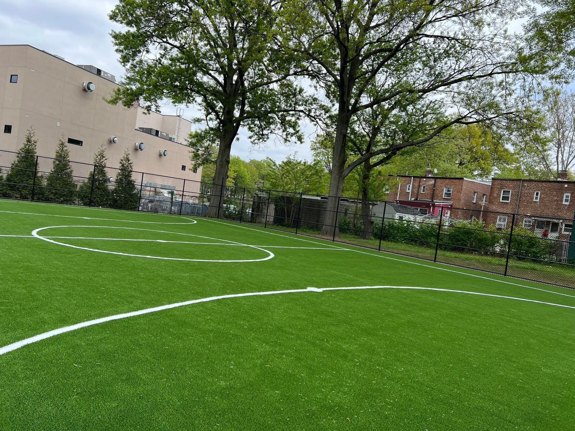 A view of a green turf field with painted white markings, flanked by trees and buildings under a cloudy sky.